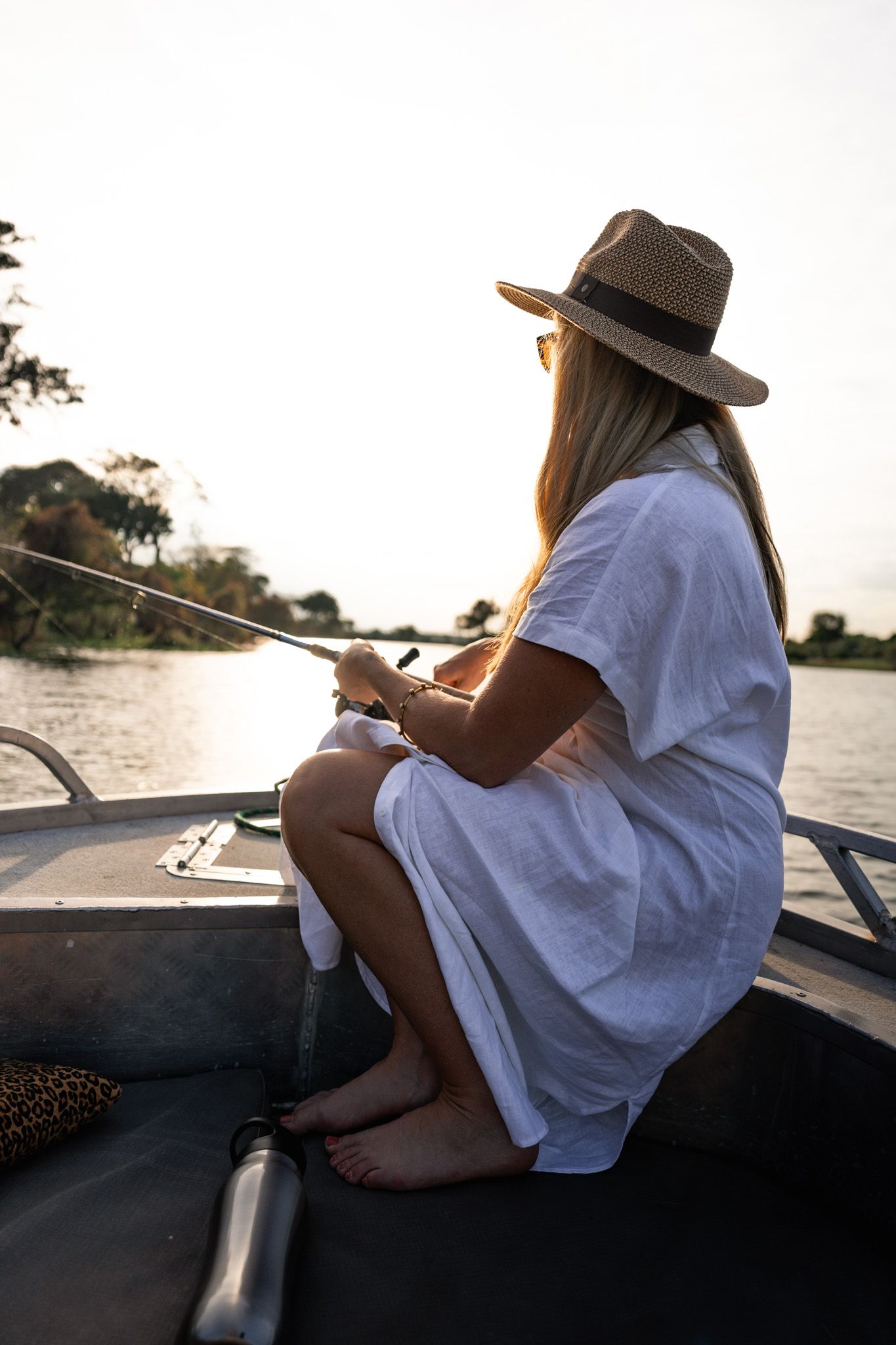 Person in white dress and hat fishing from boat during sunset on tranquil lake.