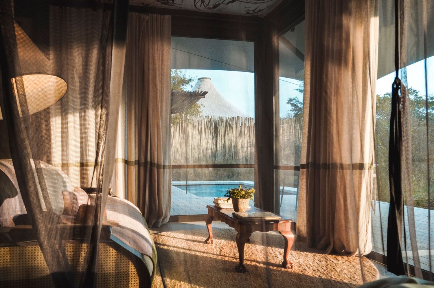 Sunlit luxury bedroom with sheer curtains, elegant furniture, and a serene pool view through large windows.