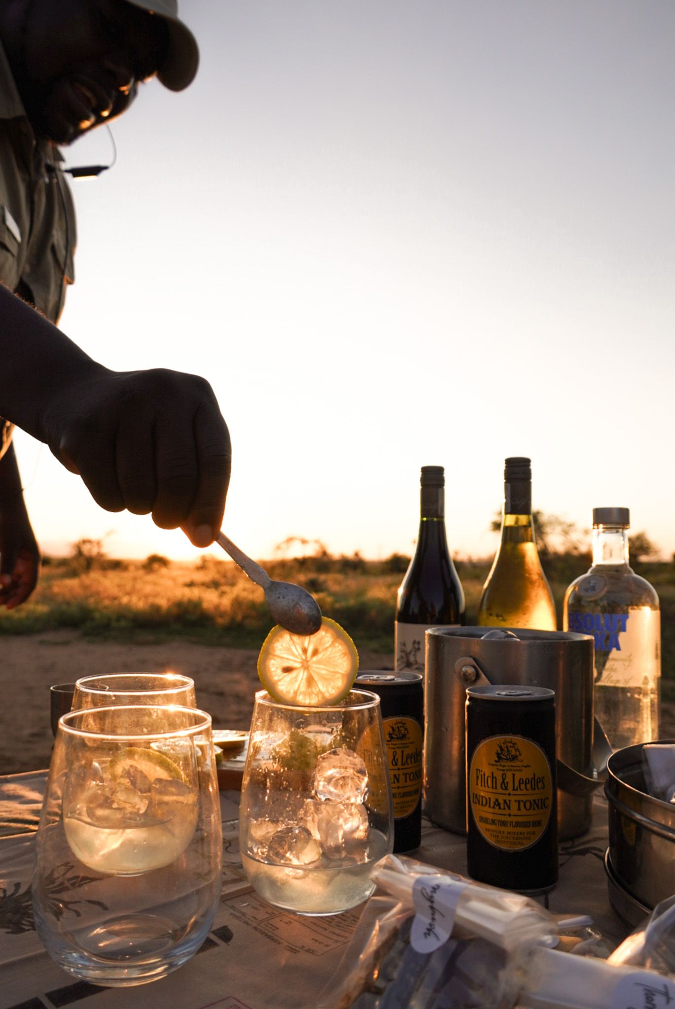 Hands pouring drinks at sunset with multiple bottles and glasses arranged on a table outdoors.