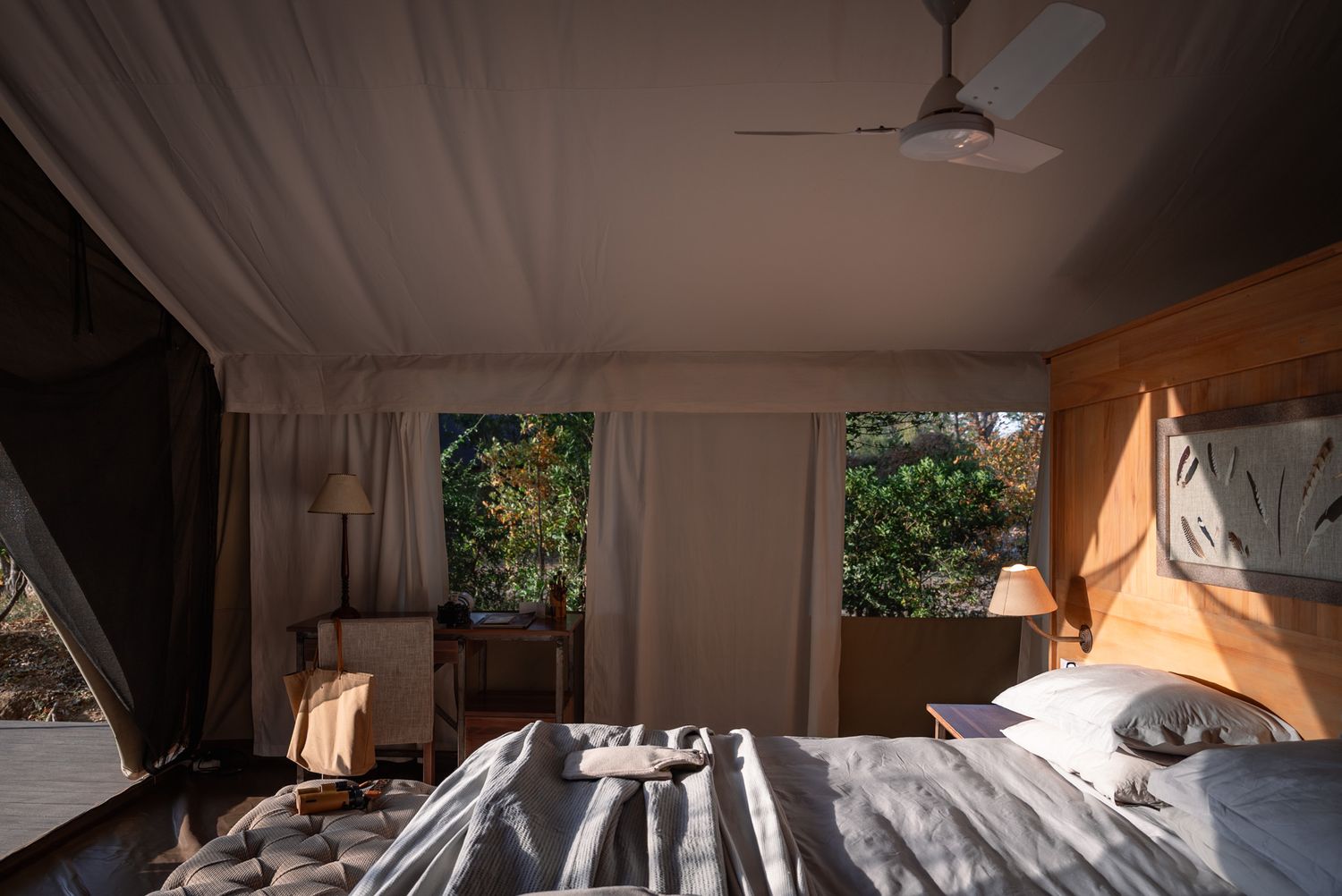 Sunlit luxury tent bedroom with ceiling fan and white drapes offering views of surrounding nature.
