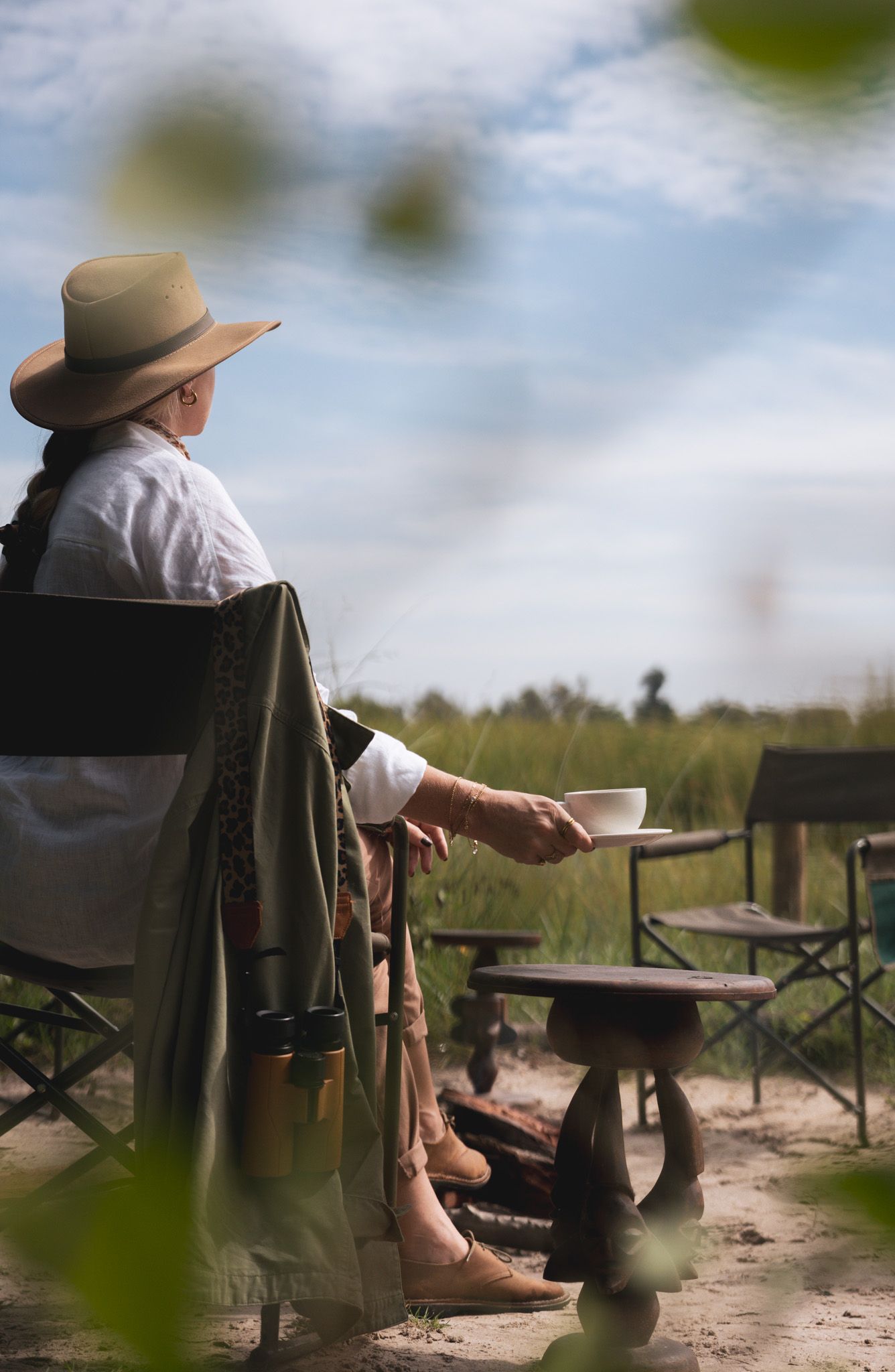 Relaxed outdoor scene with person in hat enjoying coffee in camping chair.