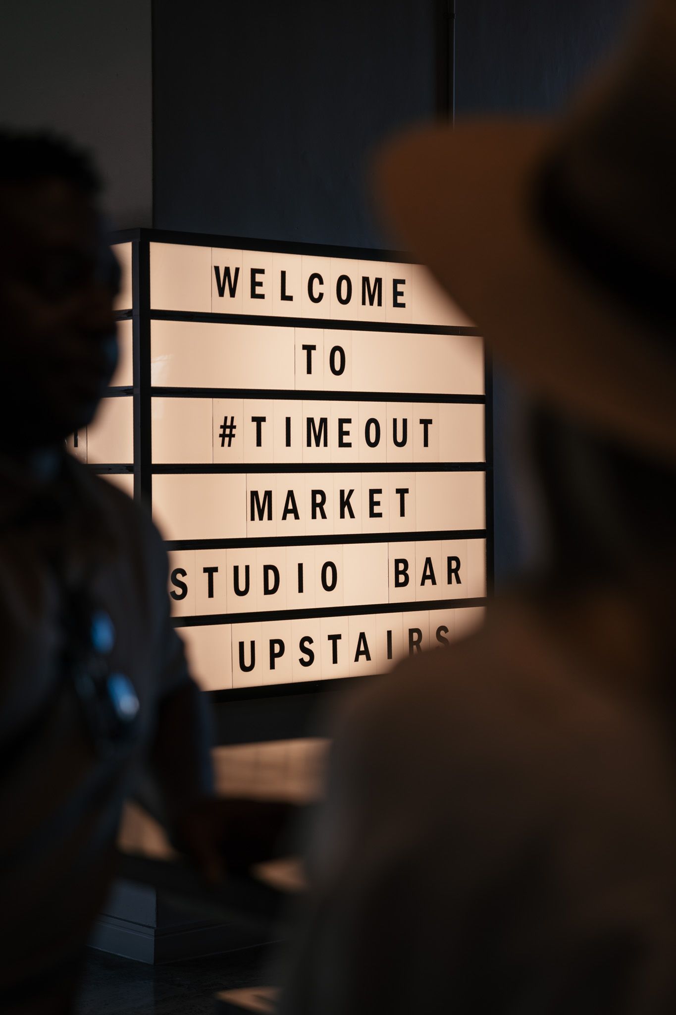 Illuminated welcome sign for Timeout Market Studio Bar with silhouettes in foreground.