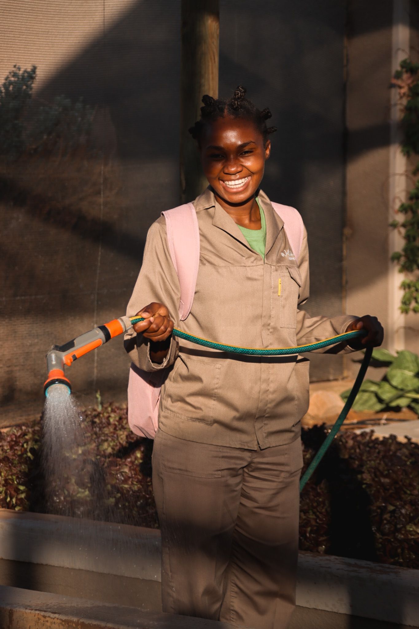 Garden worker in beige uniform waters plants with hose in evening light.