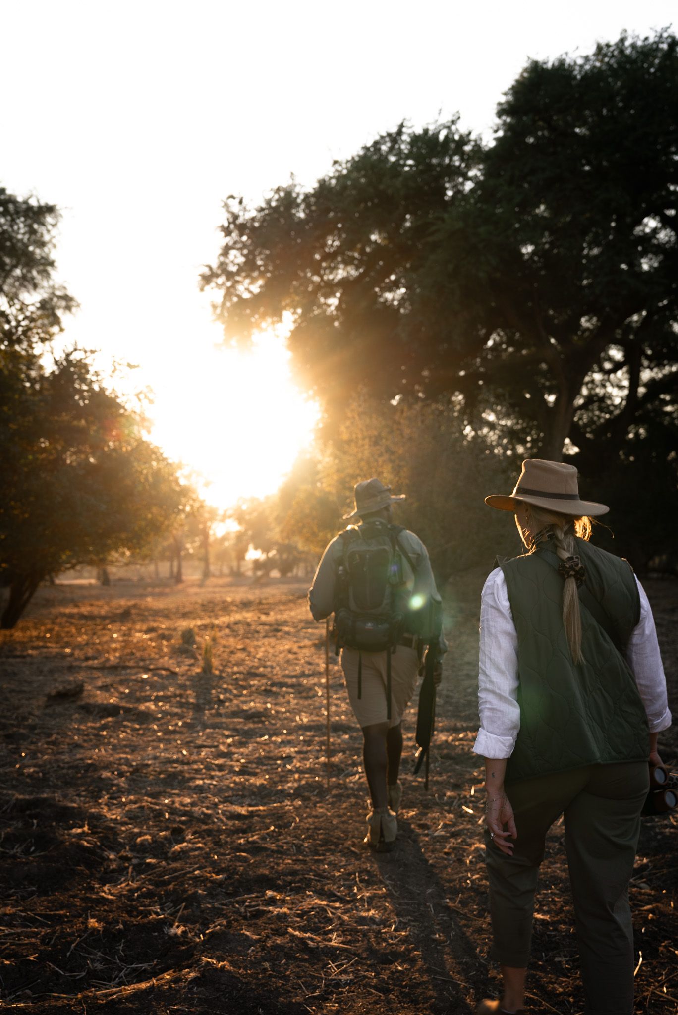 People walking on dusty trail during golden hour with sun peeking through trees.