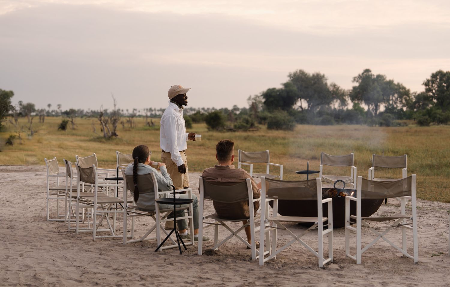 Guests seated at dining tables in a rustic outdoor setting at dusk with safari landscape in the background.