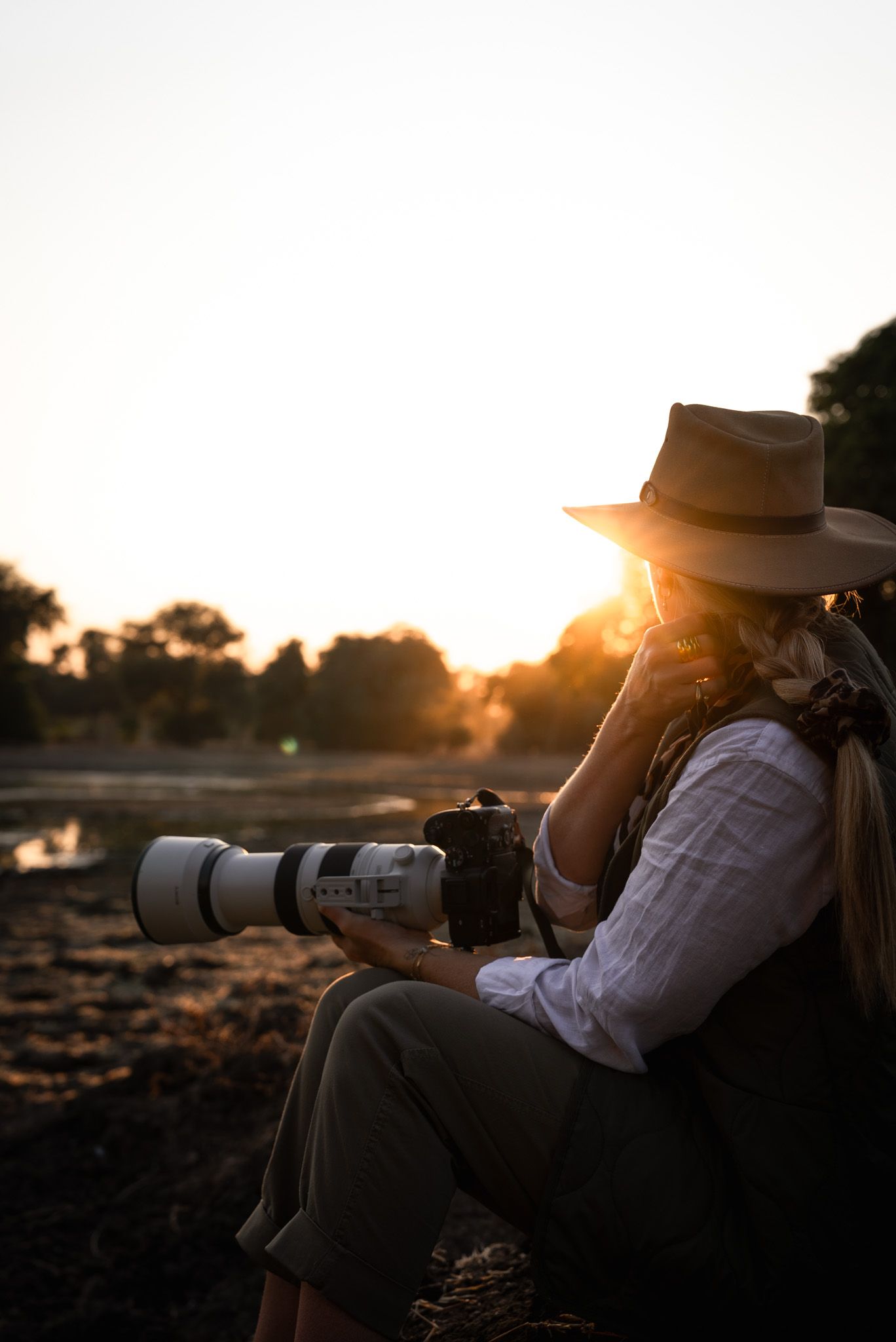 A photographer with a telephoto lens captures the golden sunset while wearing a wide-brimmed hat.