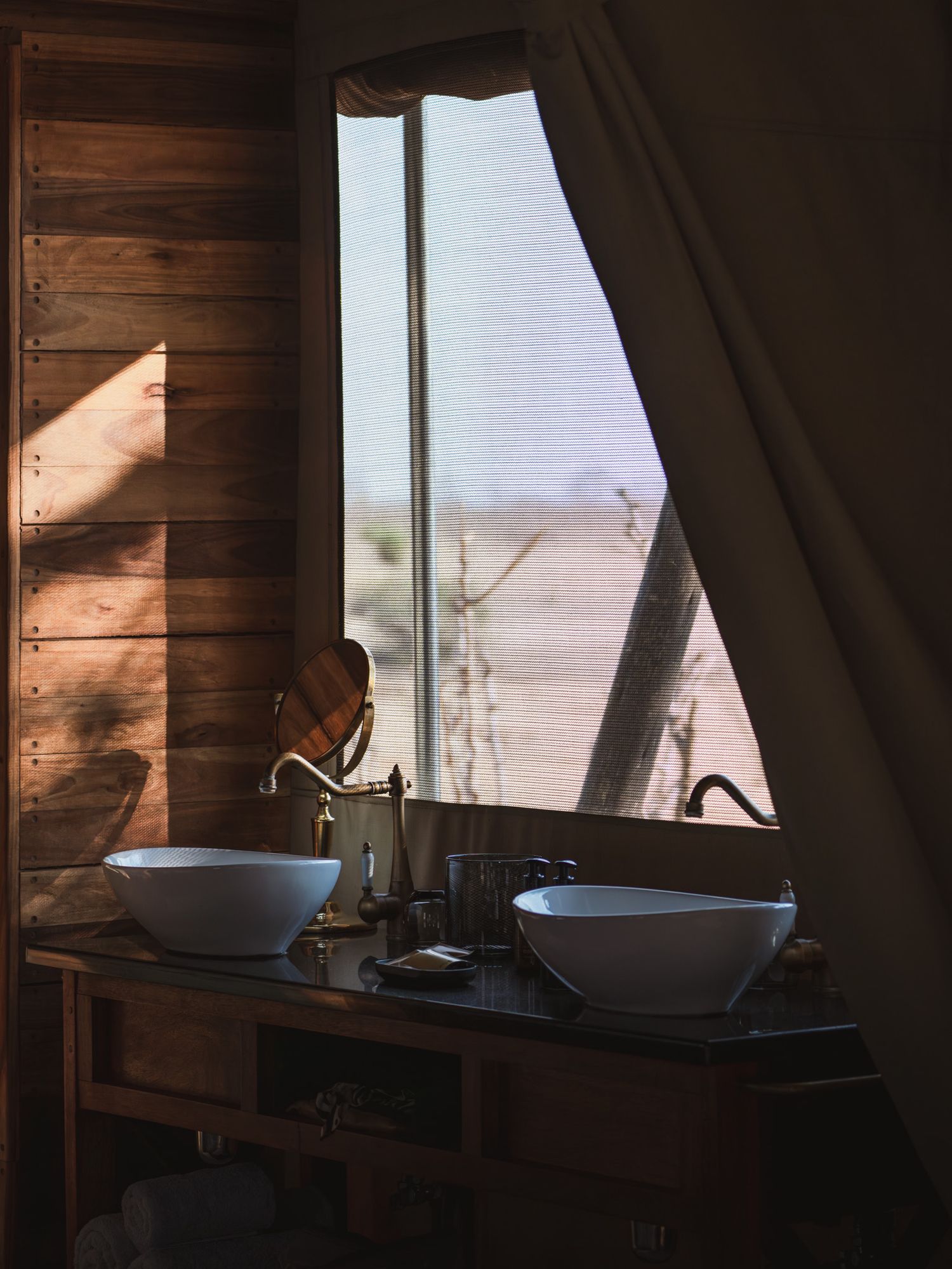 Rustic bathroom interior with wooden walls and white sinks backlit by desert landscape view.
