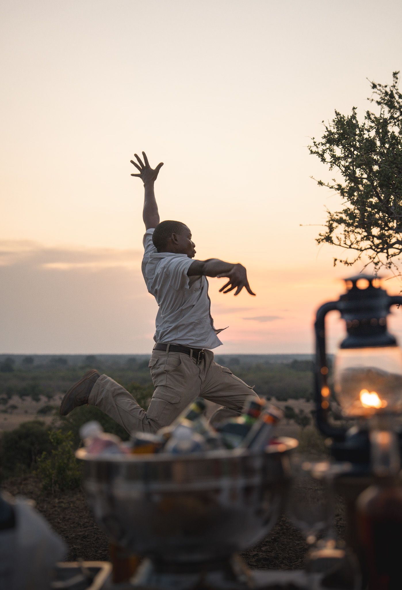 Person jumping joyfully at sunset with camping equipment and a campfire in the background.