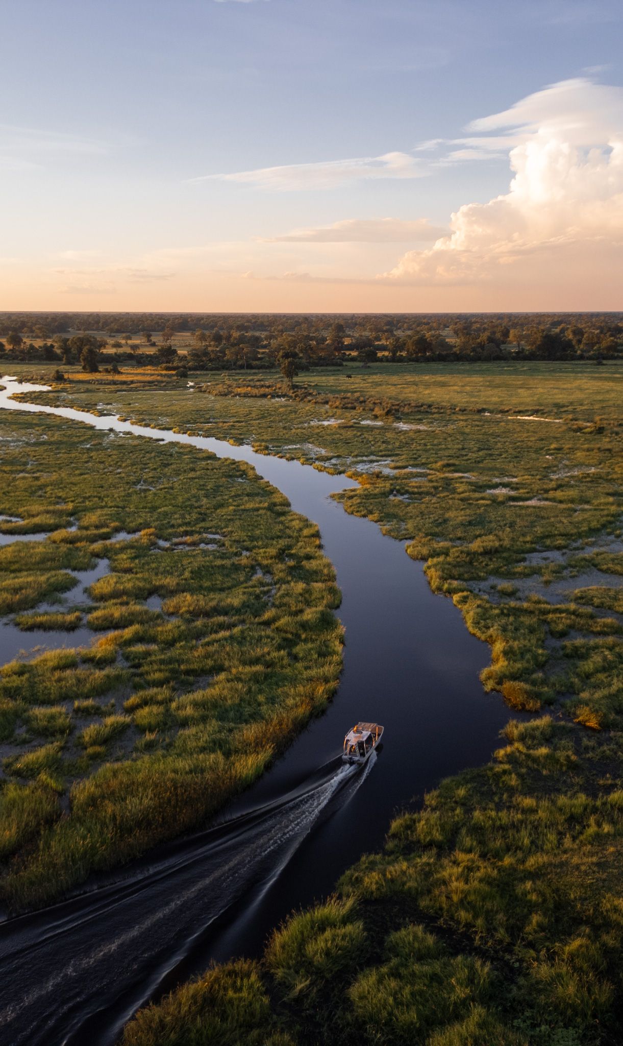 Aerial view of a boat creating wake while cruising through a winding river in marshlands at sunset.
