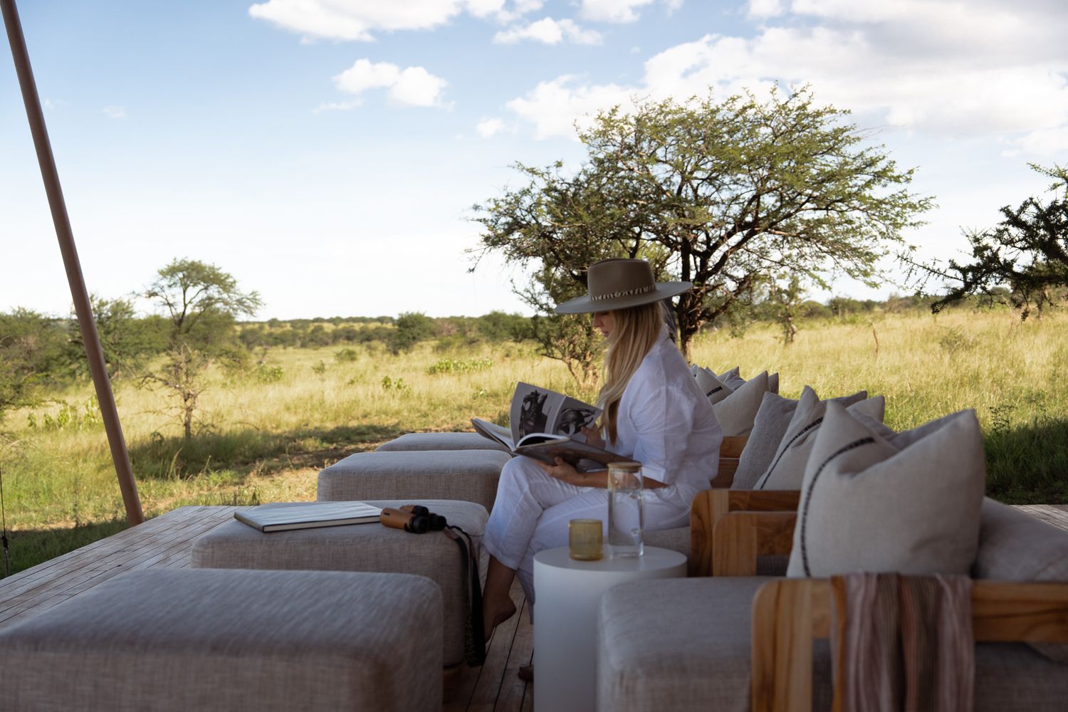 Safari-style outdoor lounge setup with neutral cushions under acacia tree in African savanna.