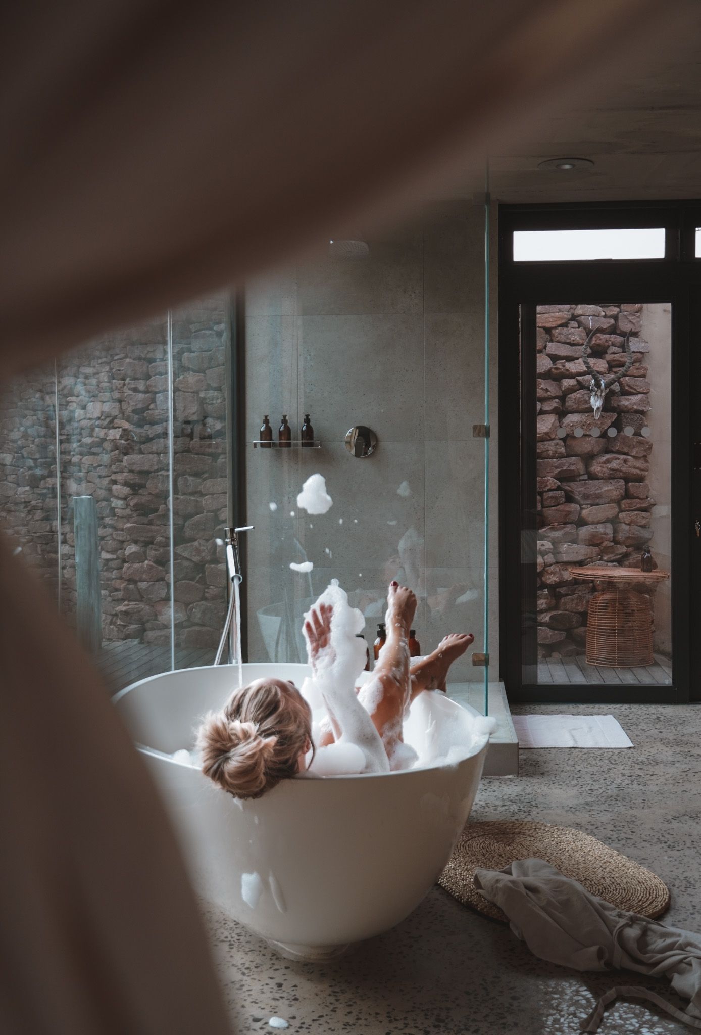 A person splashing and playing in a freestanding white bathtub set against an exposed stone wall in moody lighting.