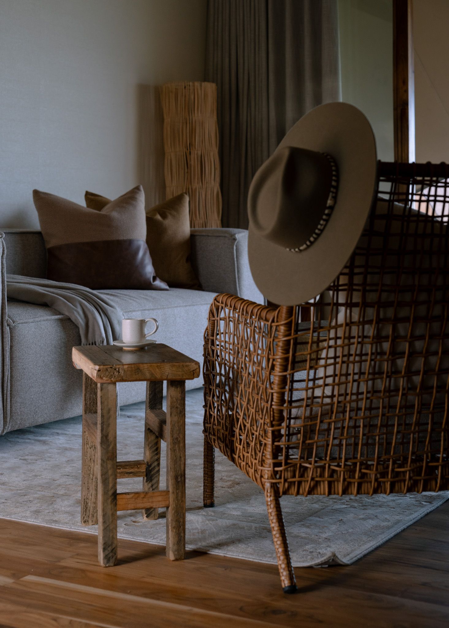 Modern living room corner with gray couch, wooden stool, and decorative hat on rattan chair.