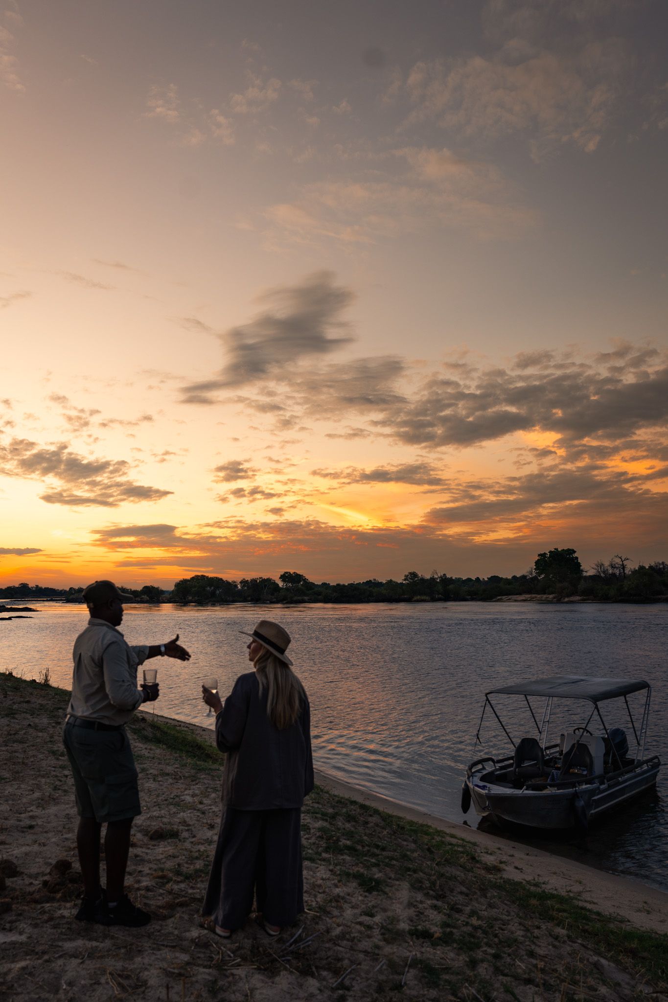 Two figures silhouetted against dramatic sunset sky by river with boat in background.