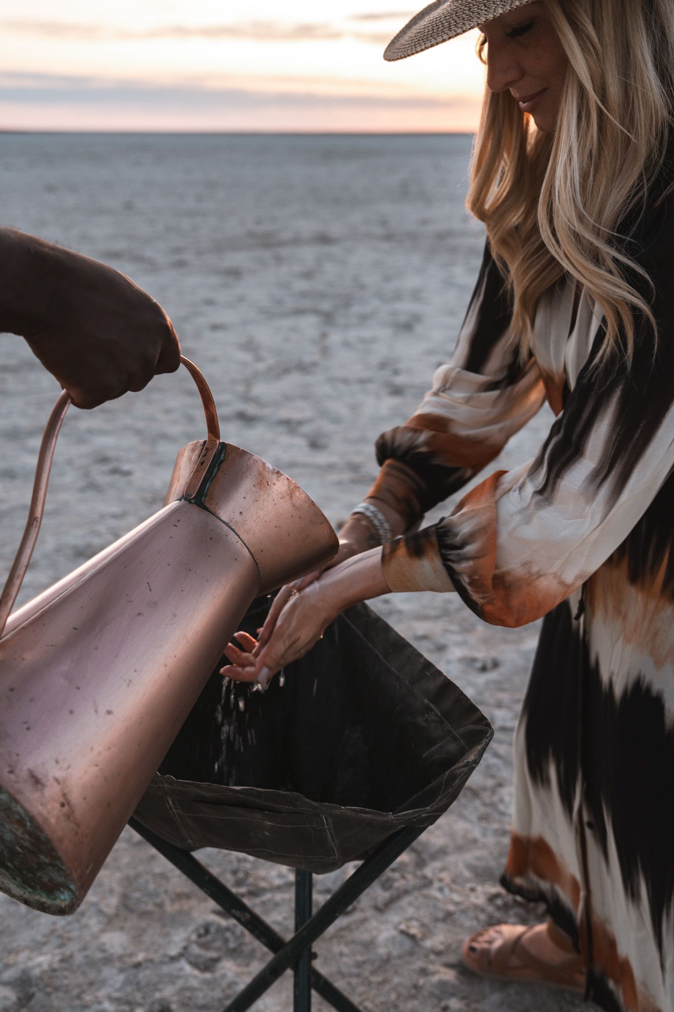 Person in hat pours water from copper kettle at desert campsite during sunset.