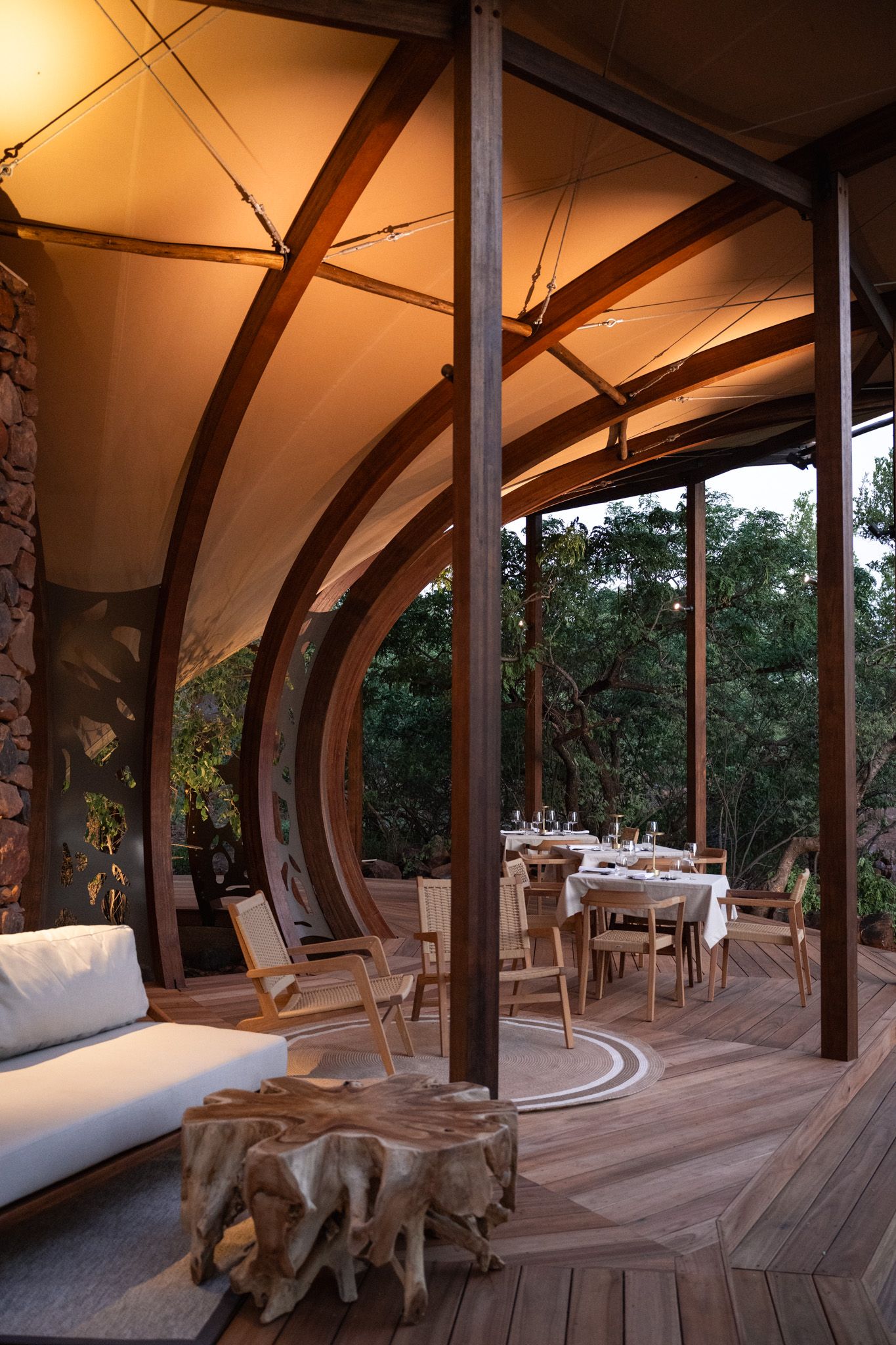 Curved wooden beams support canvas ceiling in luxury outdoor dining area at sunset.