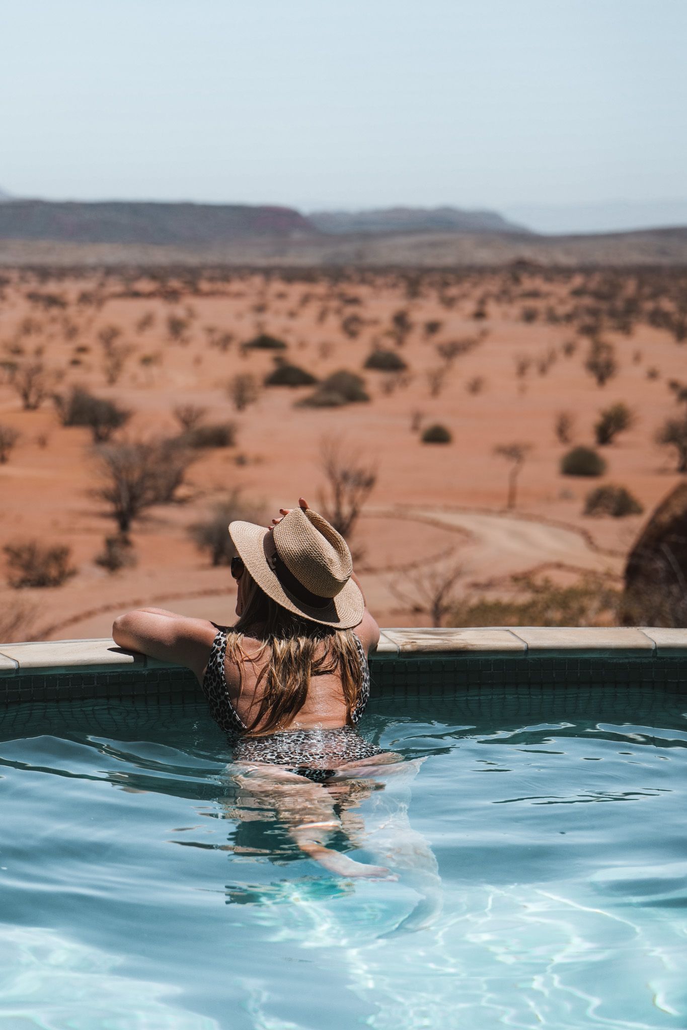 Relaxing in infinity pool overlooking desert landscape while wearing sun hat.