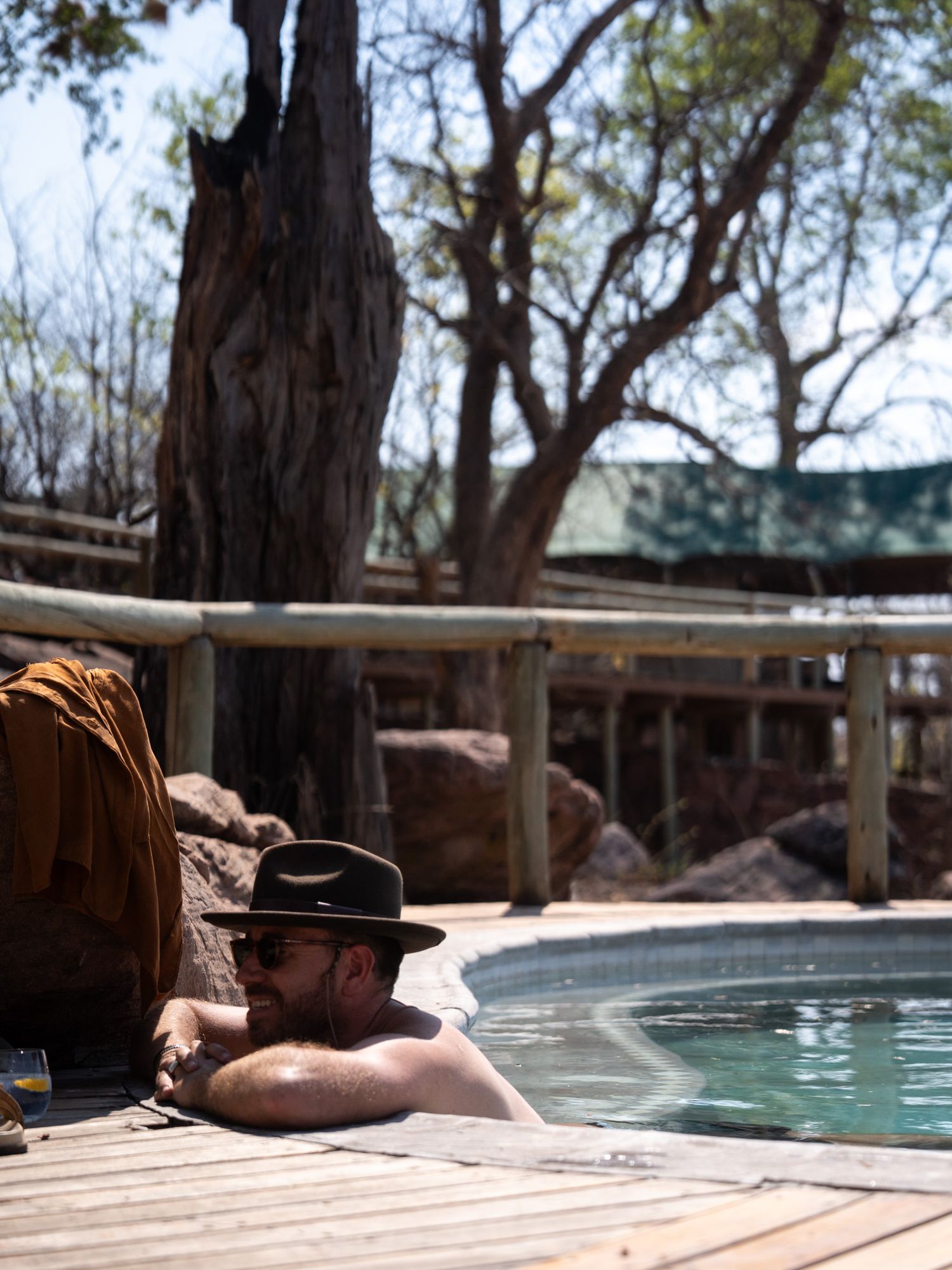 Person relaxing at the edge of a wooden deck pool surrounded by natural landscape.