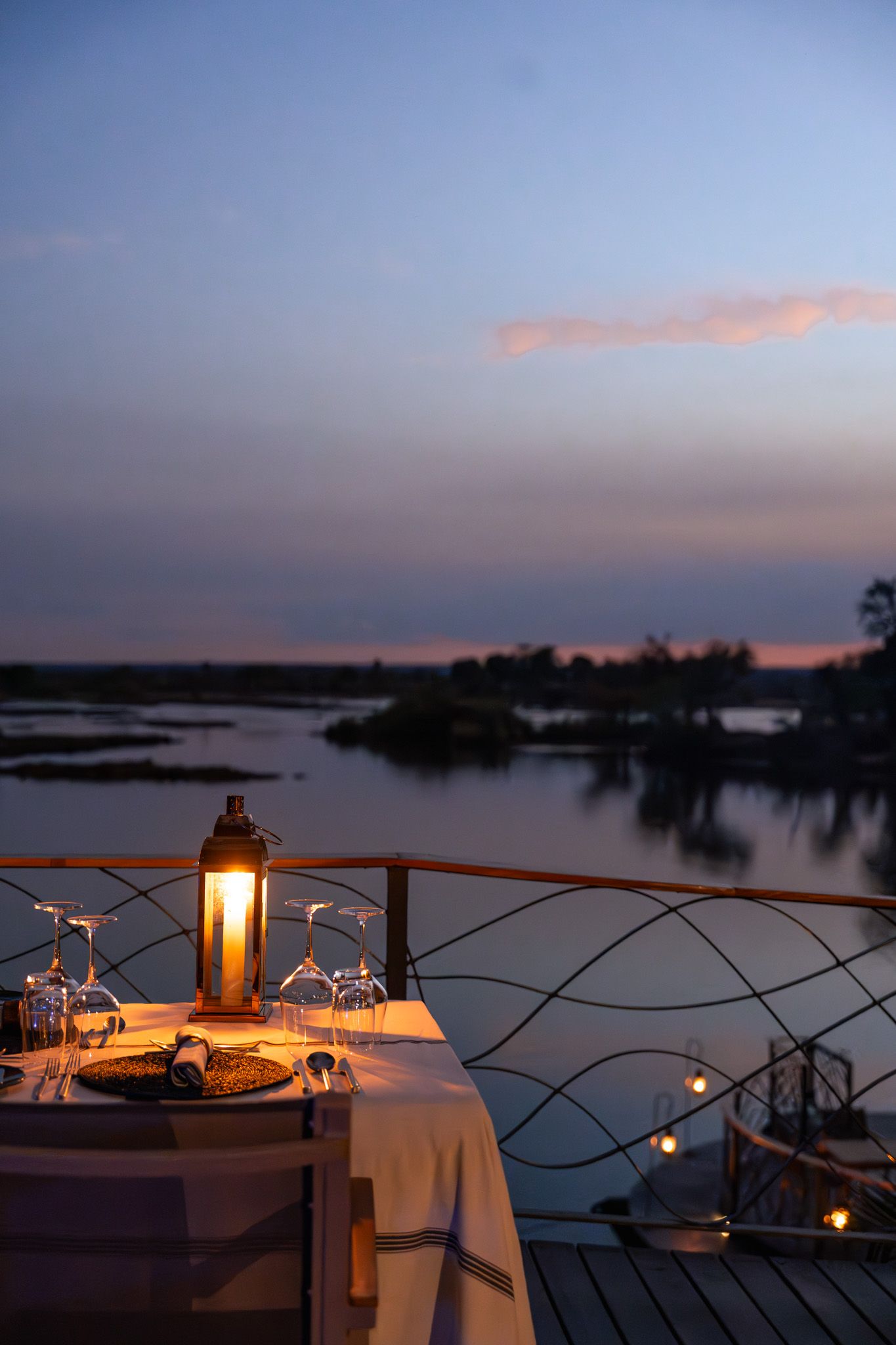 Romantic candlelit dining tables set up along a waterfront deck at sunset with glowing lanterns.