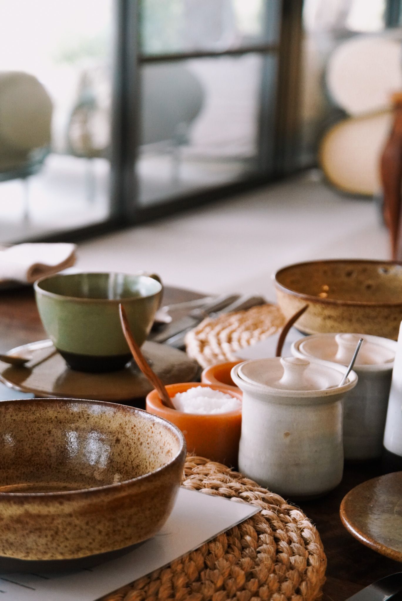 Modern table setting with earthy ceramic bowls and cups arranged on woven rattan placemats.