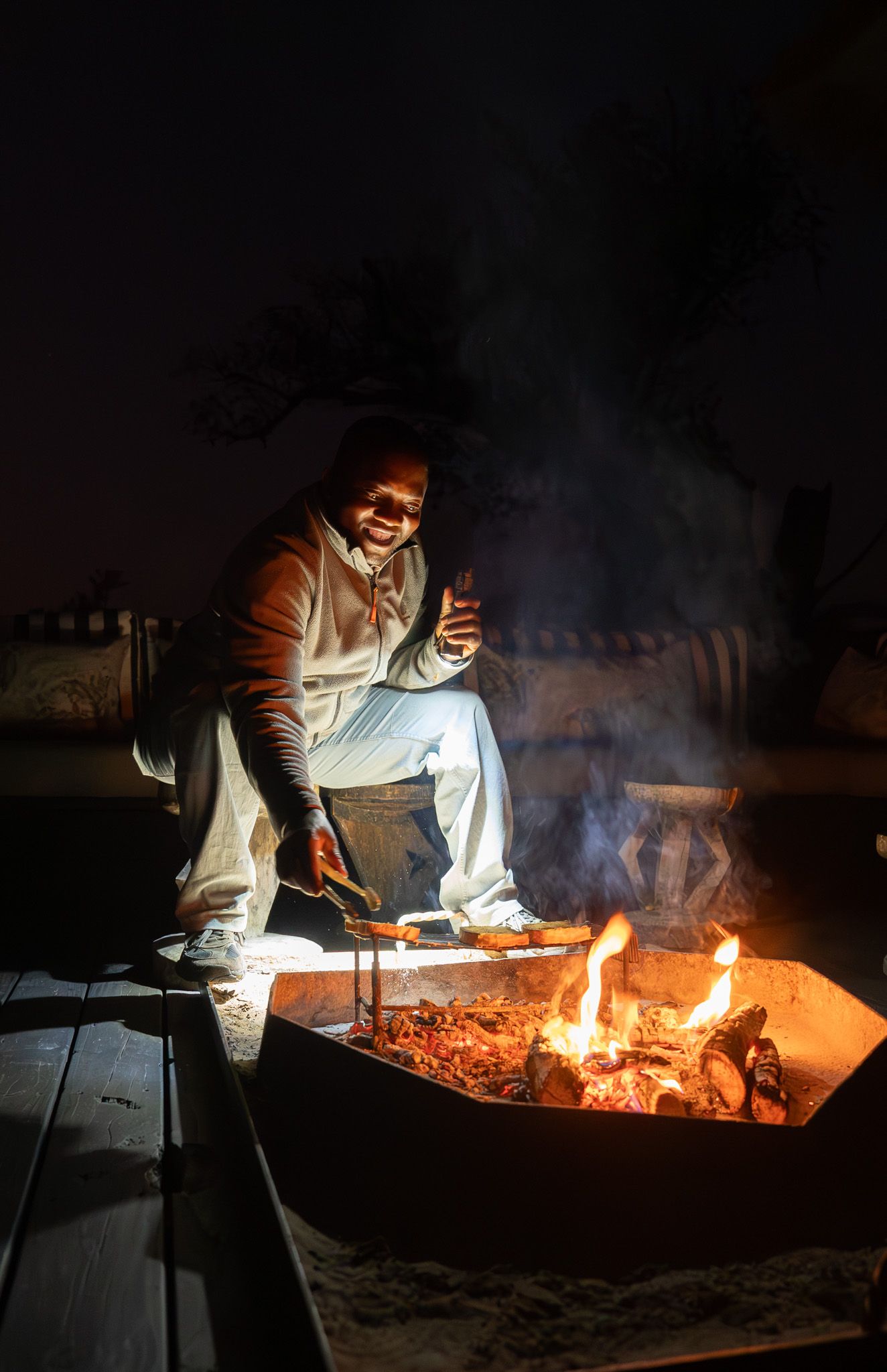 Person warming by outdoor fire pit at night with smoke rising into darkness.