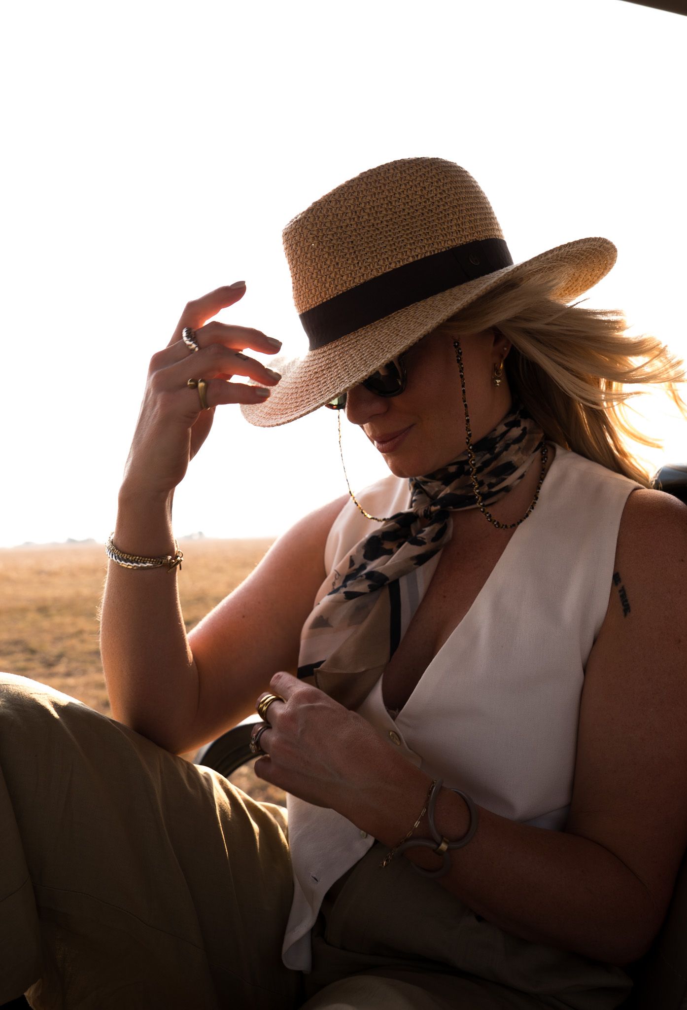 An elegant safari-goer adjusts their hat while backlit by the setting sun.