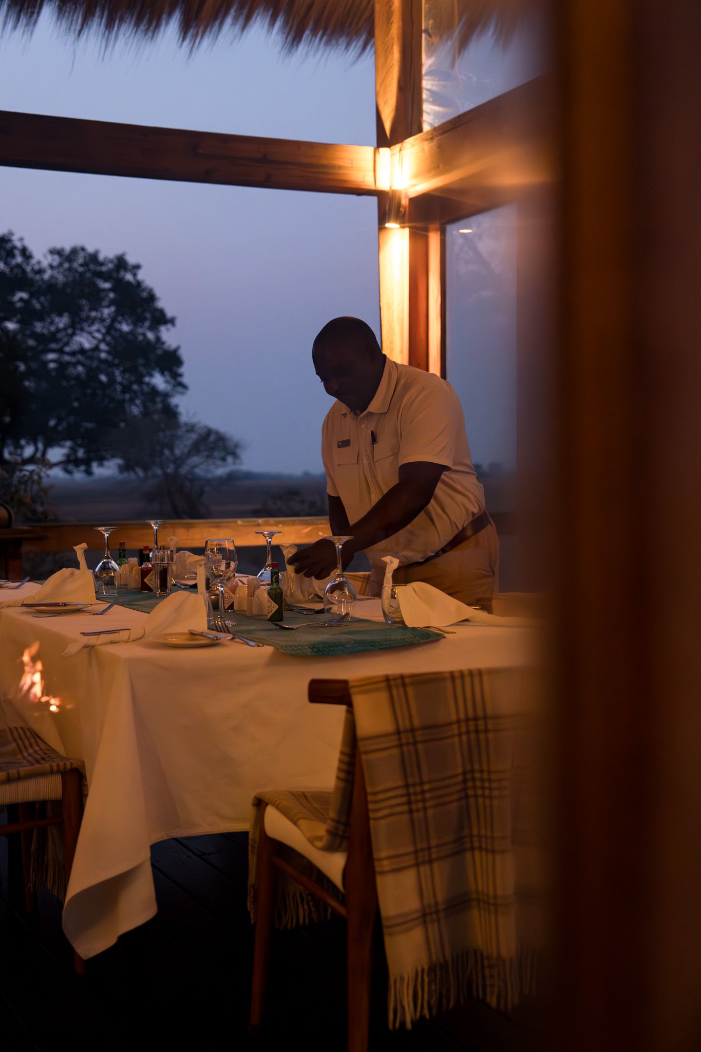 Server setting an elegant dinner table at dusk under a thatched roof.