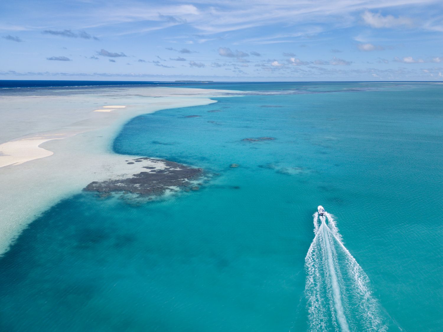 Aerial view of a boat leaving a white wake trail through turquoise ocean waters near a coral reef.