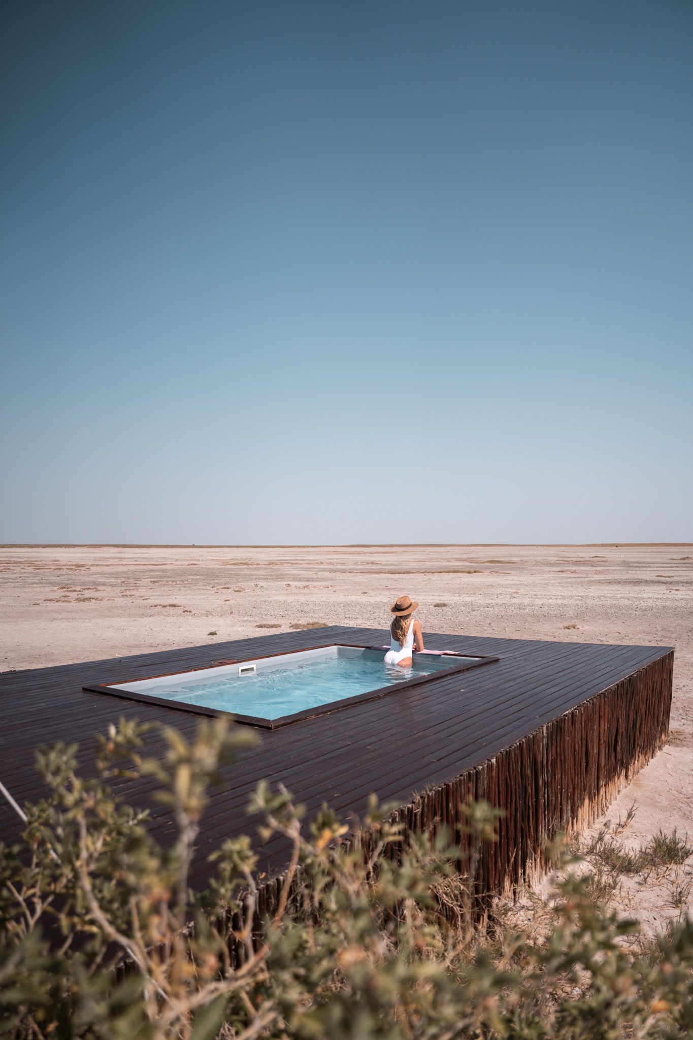 Modern plunge pool on wooden deck overlooking vast desert landscape under clear blue sky.
