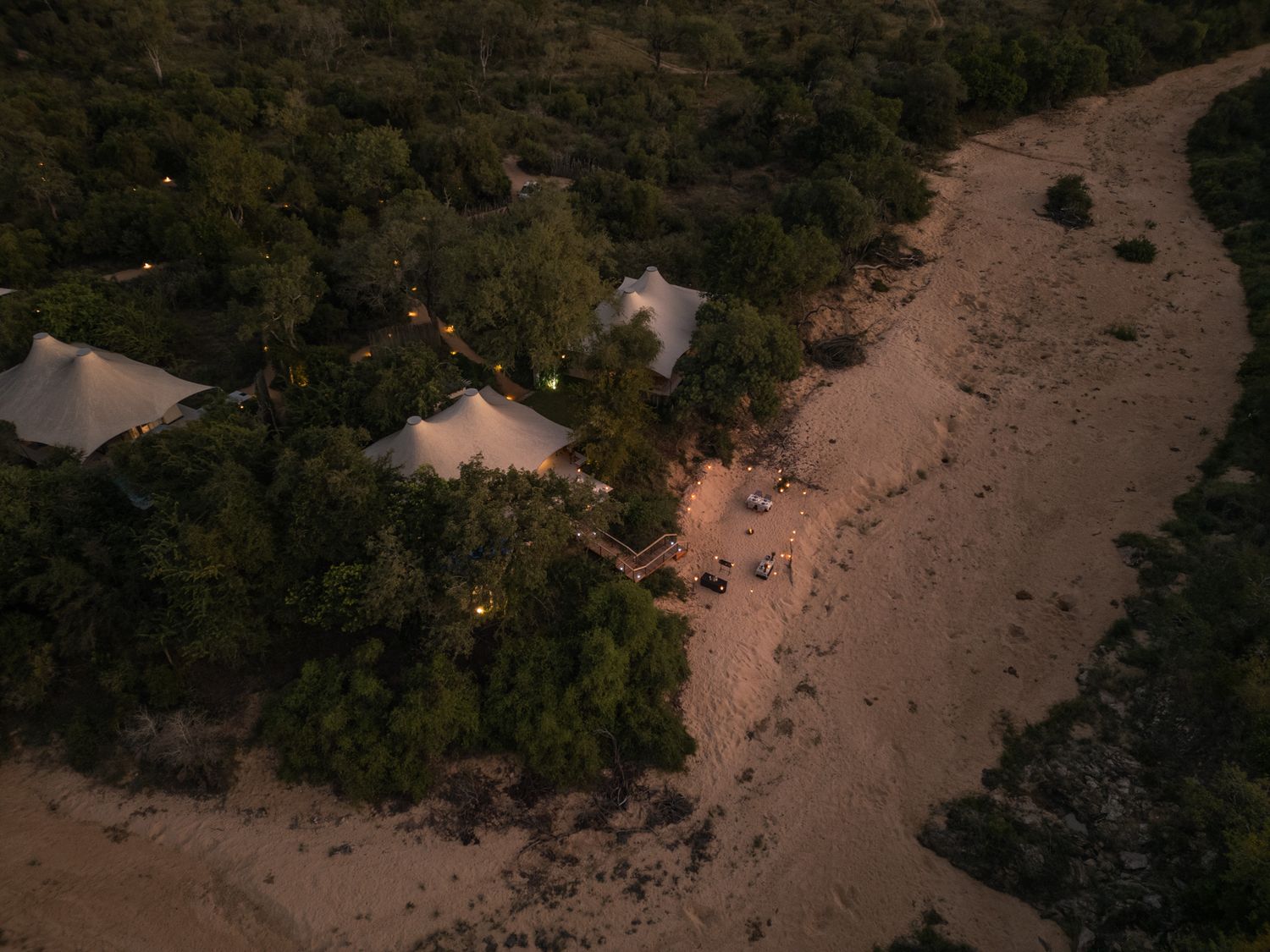 Night aerial view of glamping tents nestled among trees along a sandy path.