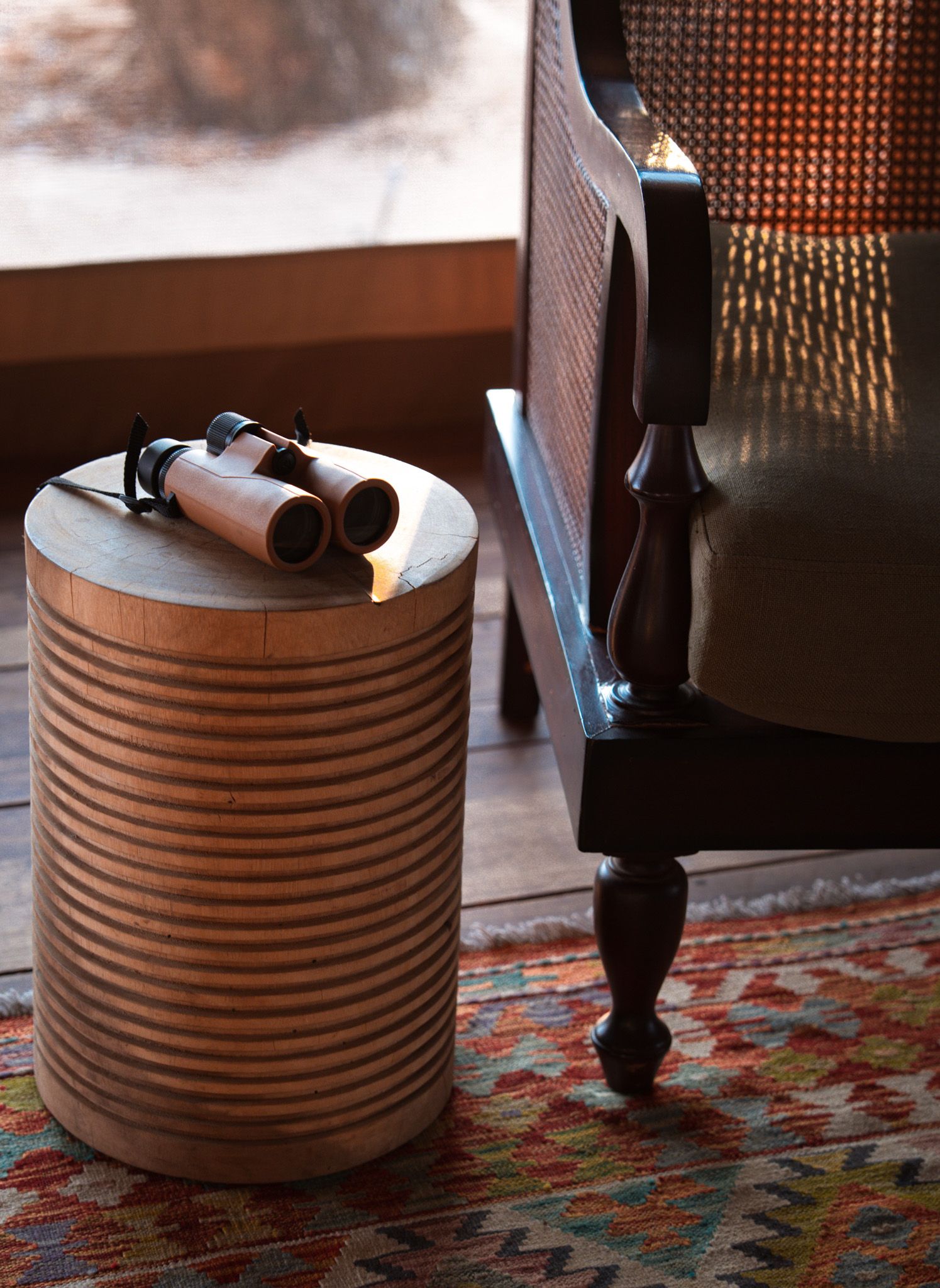 Vintage binoculars rest on a cylindrical metal drum side table next to a wooden chair.