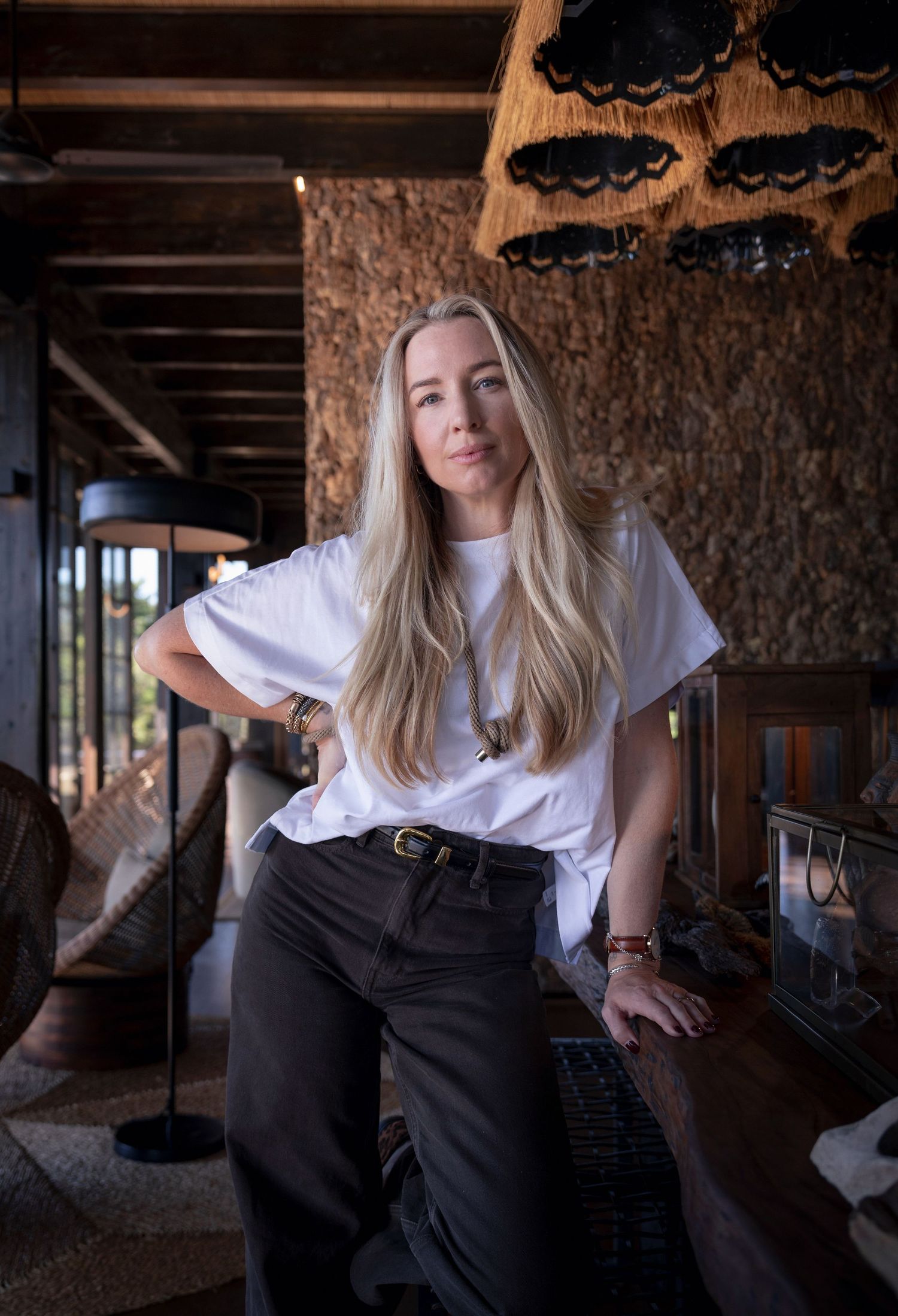 Blonde woman in white t-shirt and dark pants poses confidently in a rustic interior setting.