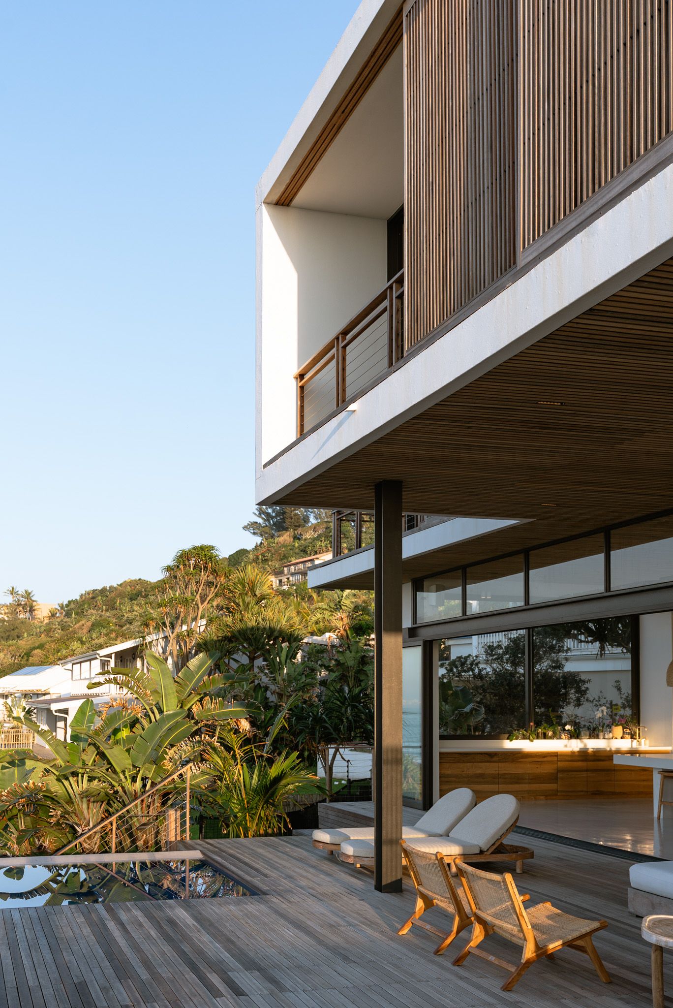 Modern coastal home exterior with wooden deck, lounge chairs, and tropical landscaping.