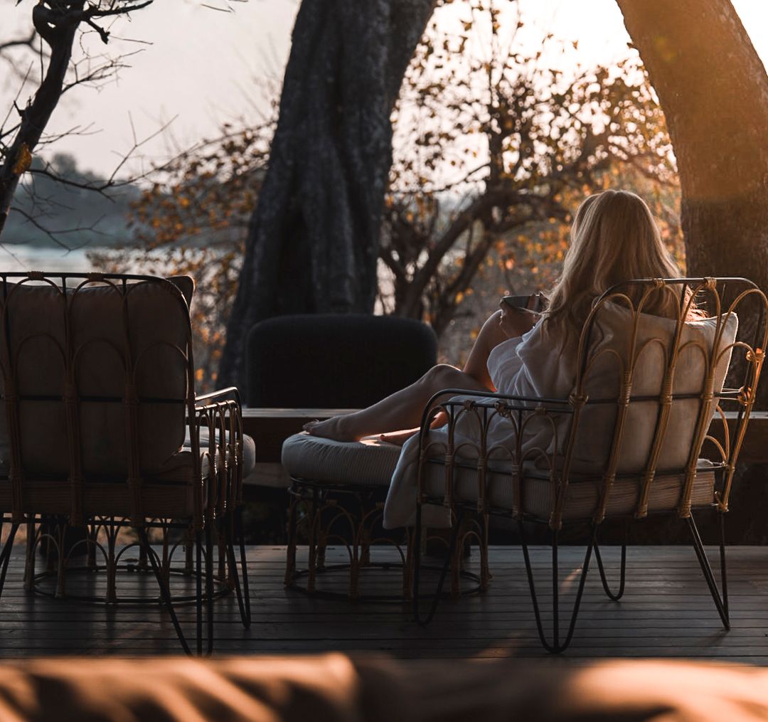 Woman relaxing in wicker chair on wooden deck at sunset, holding drink, surrounded by trees in safari lodge setting.