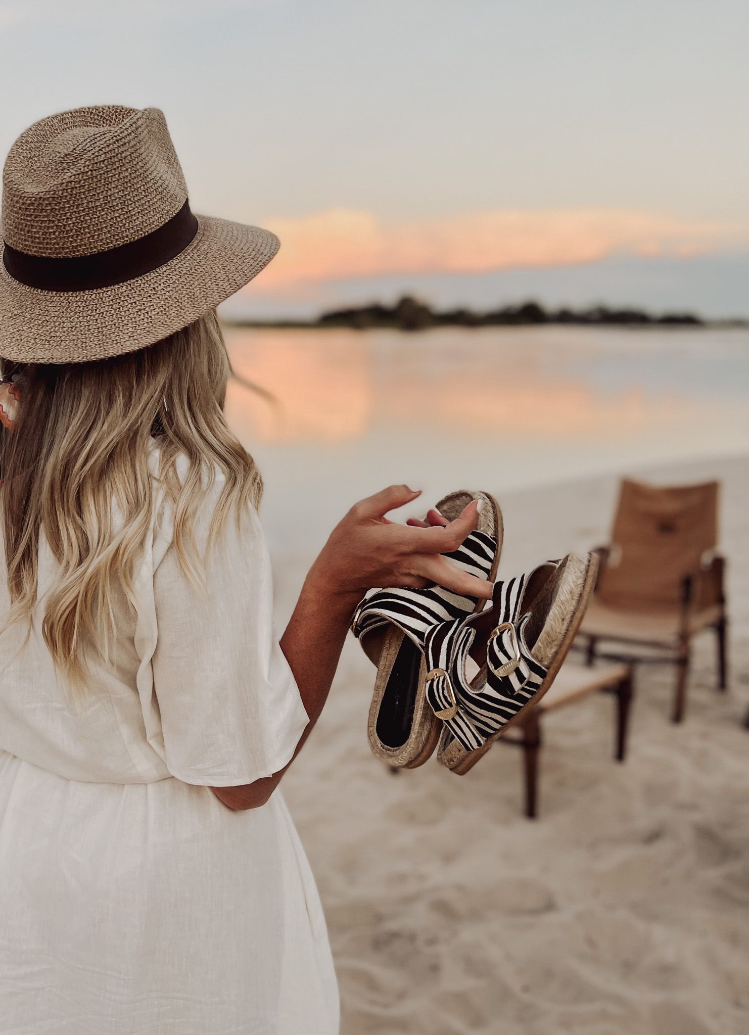 Beach scene with straw hat and striped shoes against sunset water view.