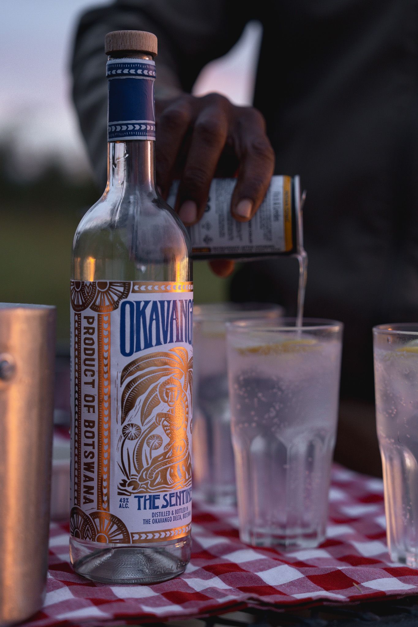 Bottle of Okavango spirit on checkered tablecloth with glasses at sunset.