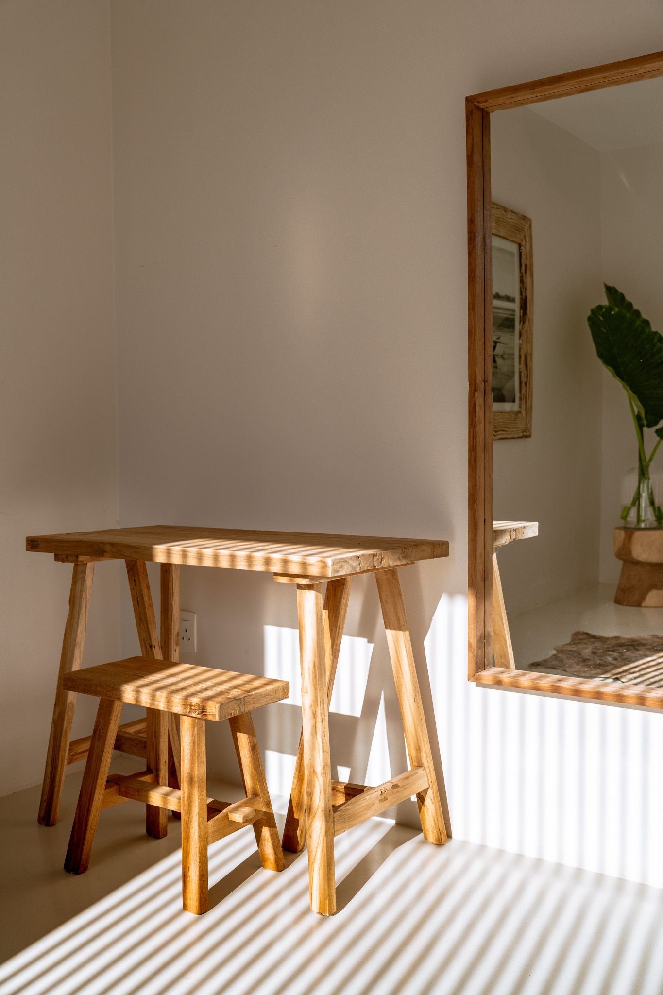 Minimalist wooden stools and table casting dramatic shadows in a sunlit white room with mirror.