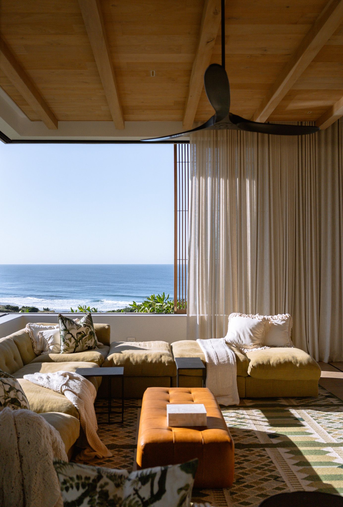 Sunlit coastal living room with comfortable sectional sofa and ocean view through floor-to-ceiling windows.
