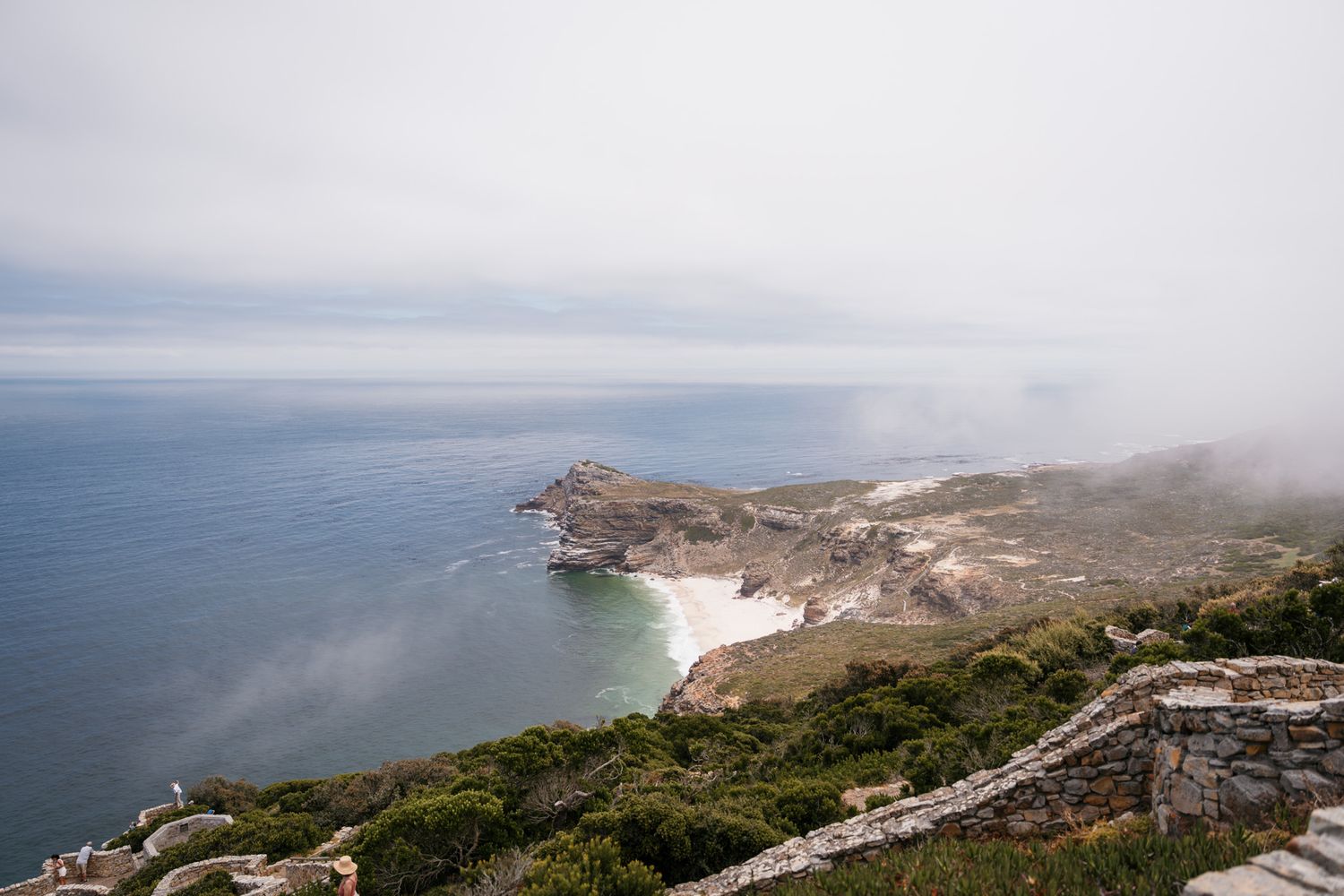 Foggy coastal cliffs with white sand beach and turquoise waters below.