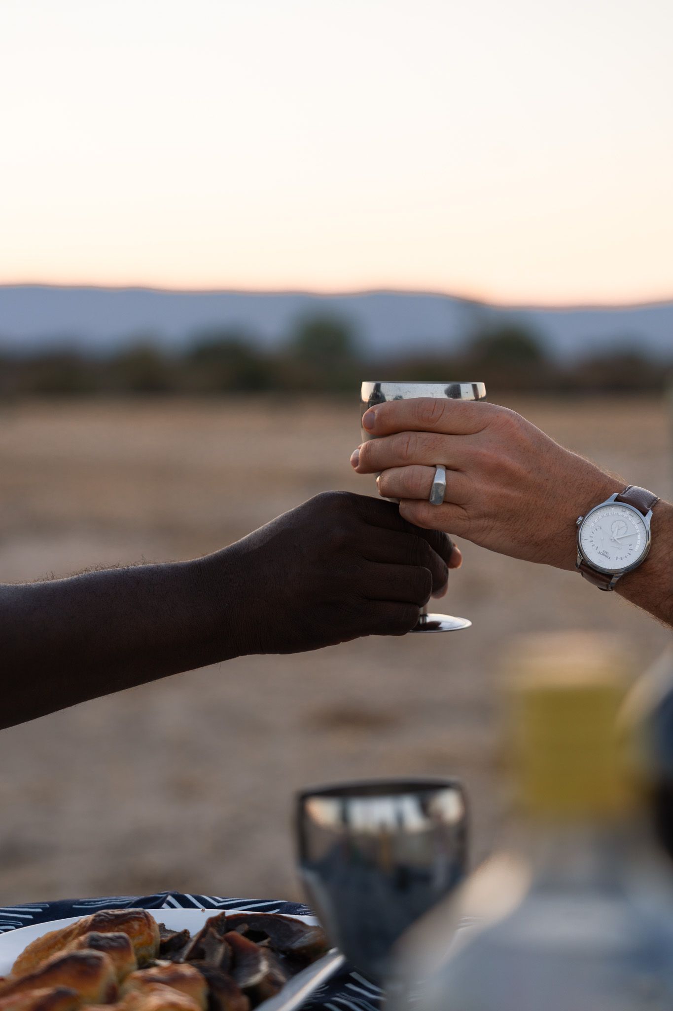 Hands exchanging objects at sunset with mountains in the background.
