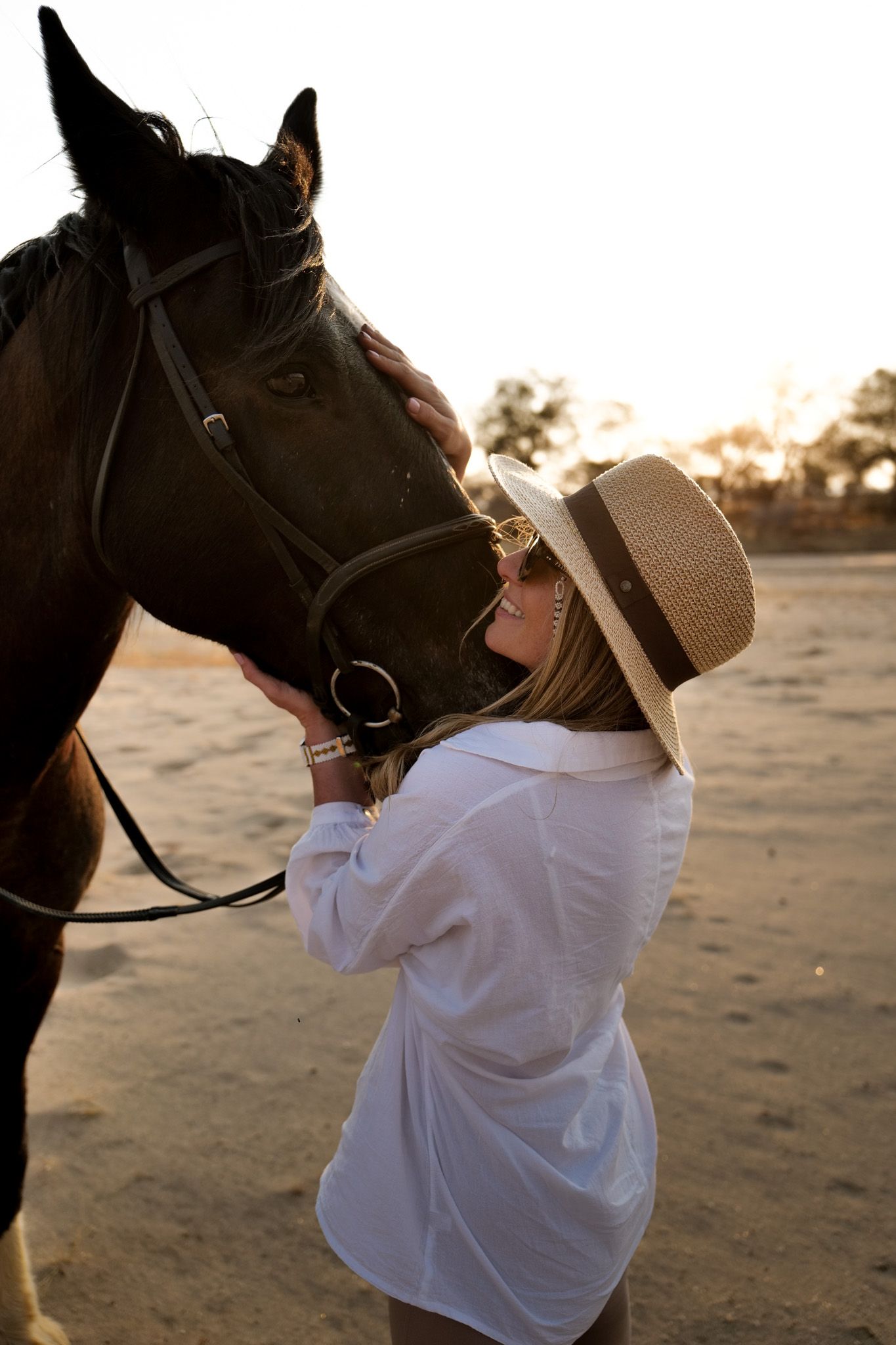 Person in white shirt and hat bonding with dark horse during golden hour.