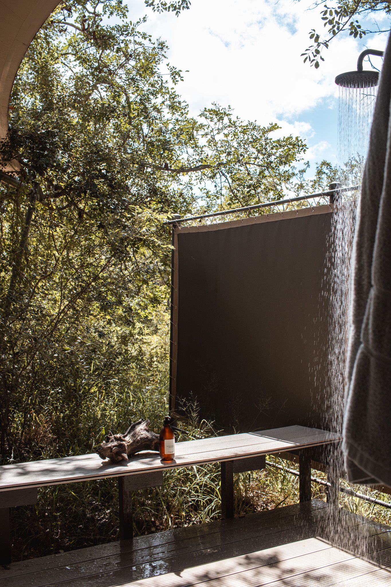 Outdoor shower with wooden bench surrounded by lush greenery and natural sunlight streaming through the trees.
