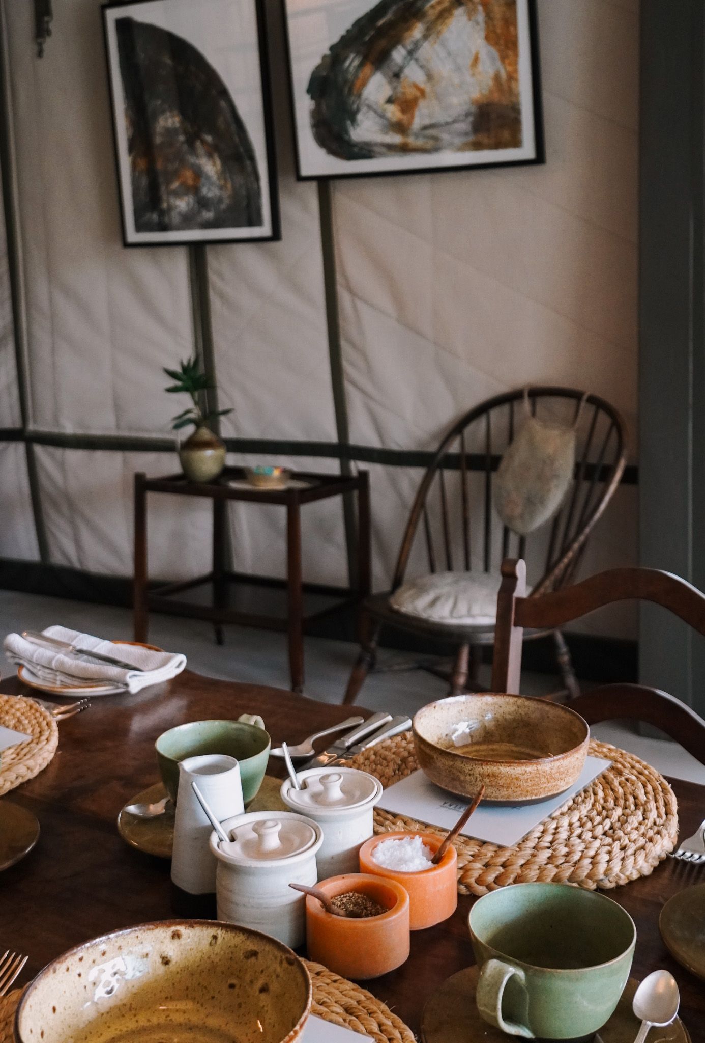 Cozy dining setup with wooden chairs, artwork, and colorful ceramic mugs on woven placemats.