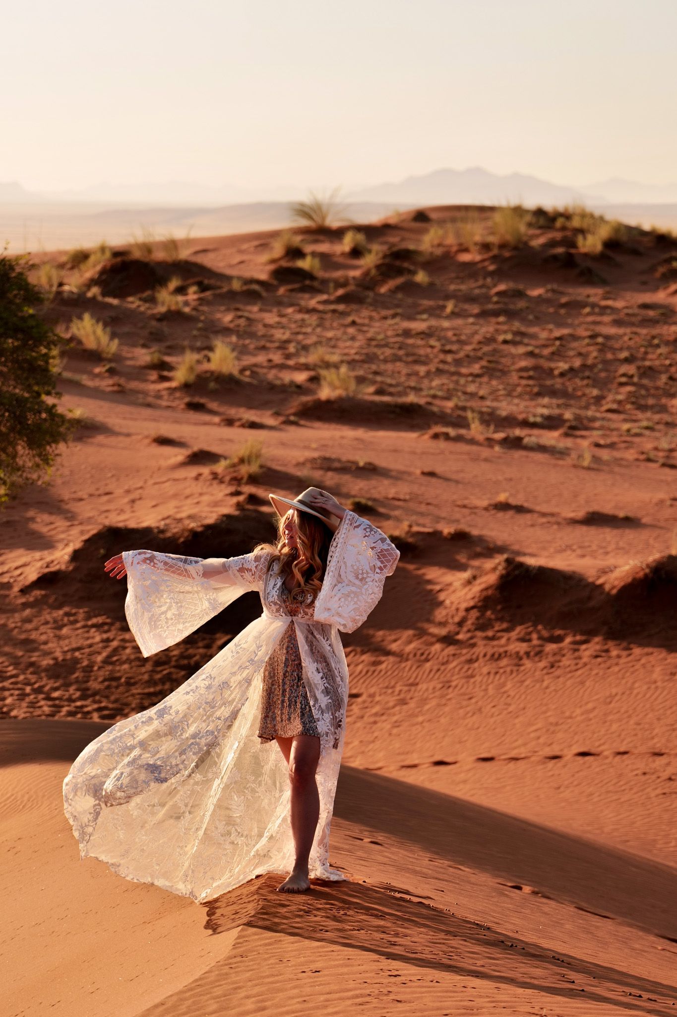 Flowing white dress catches wind against red desert sand dunes at sunset.