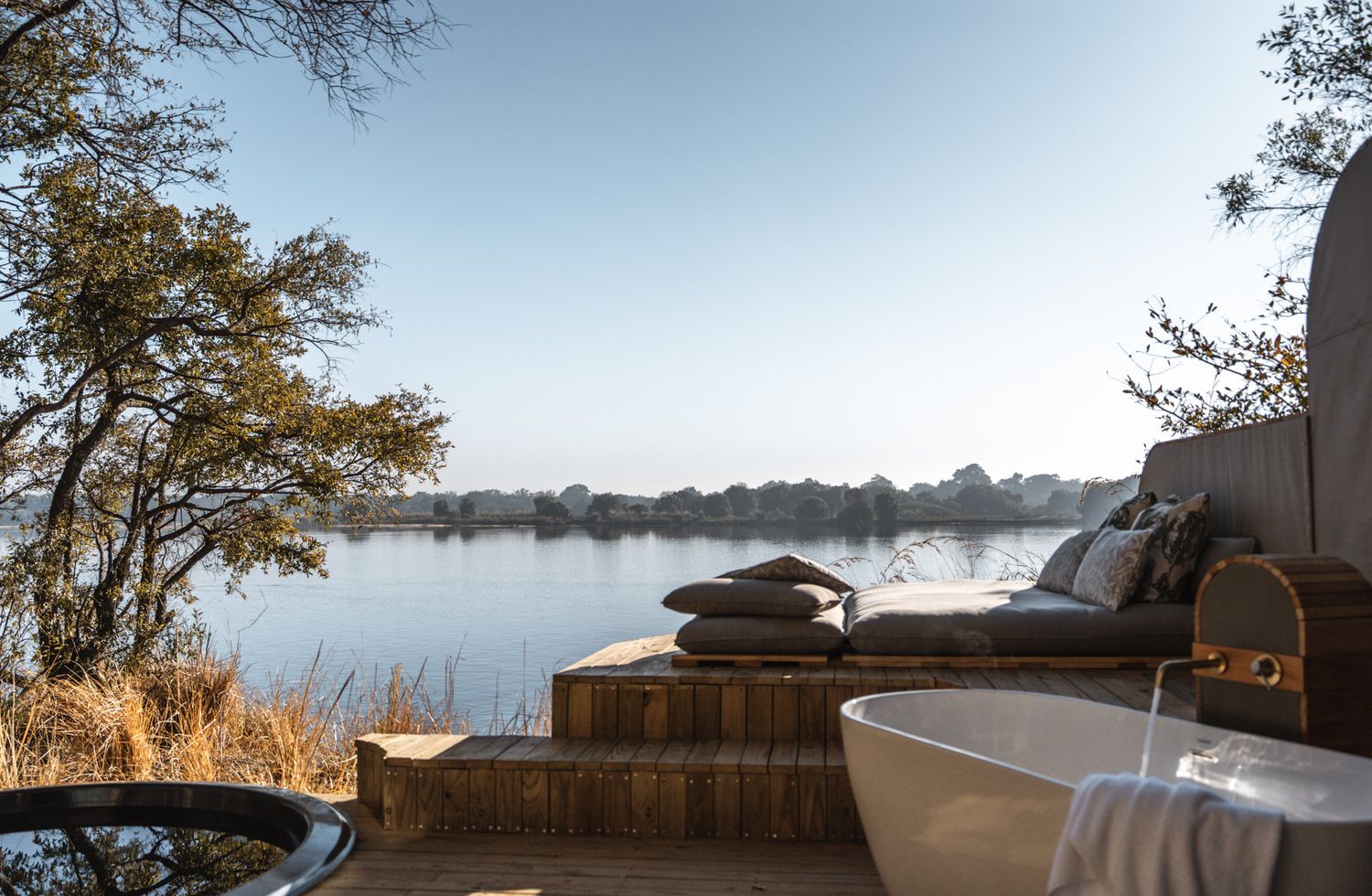 Freestanding bathtub on wooden deck overlooking peaceful lake waters at sunrise.