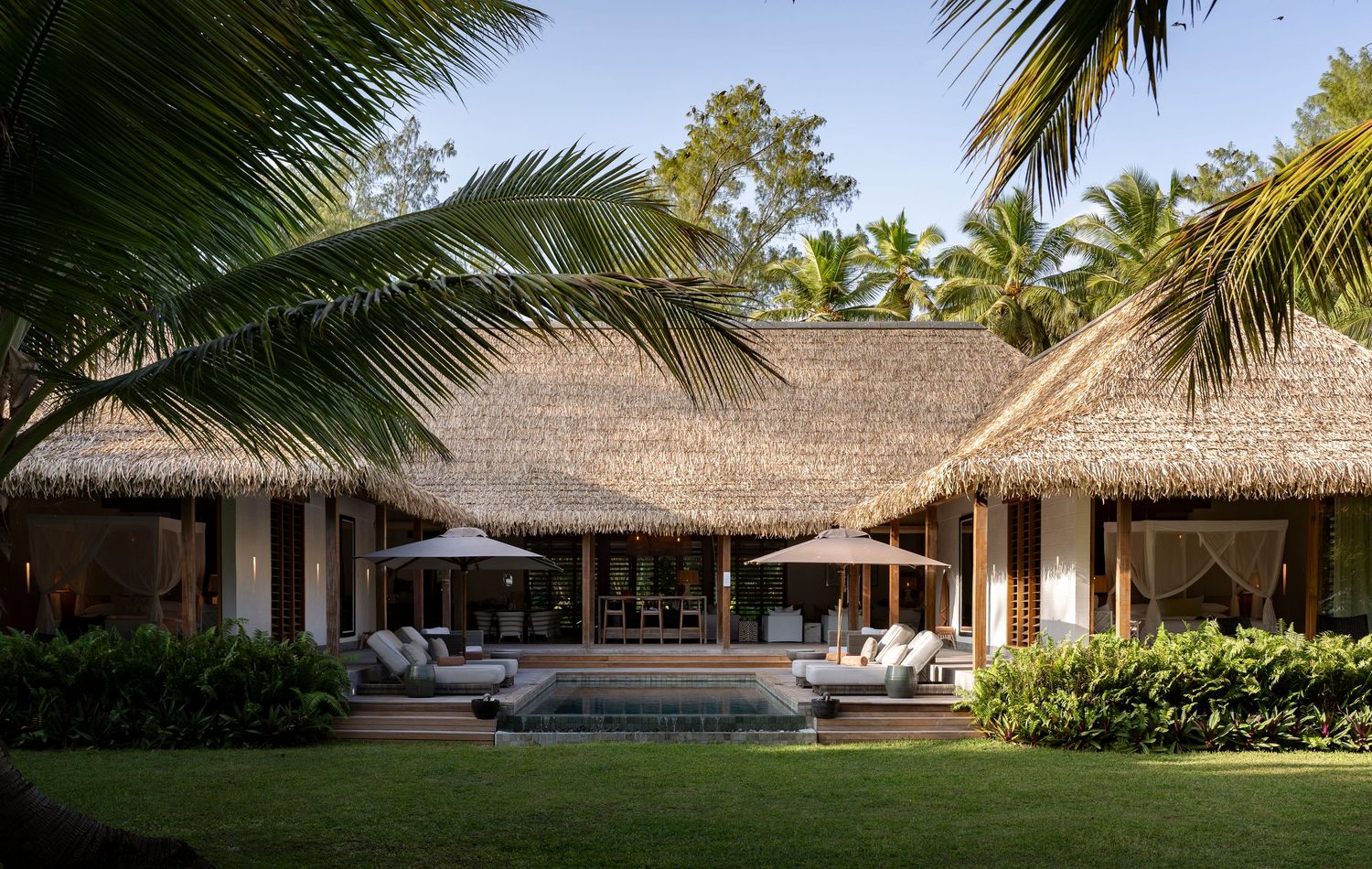 Luxury tropical villa with thatched roof, outdoor lounge chairs, pool, and lush palm tree surroundings.