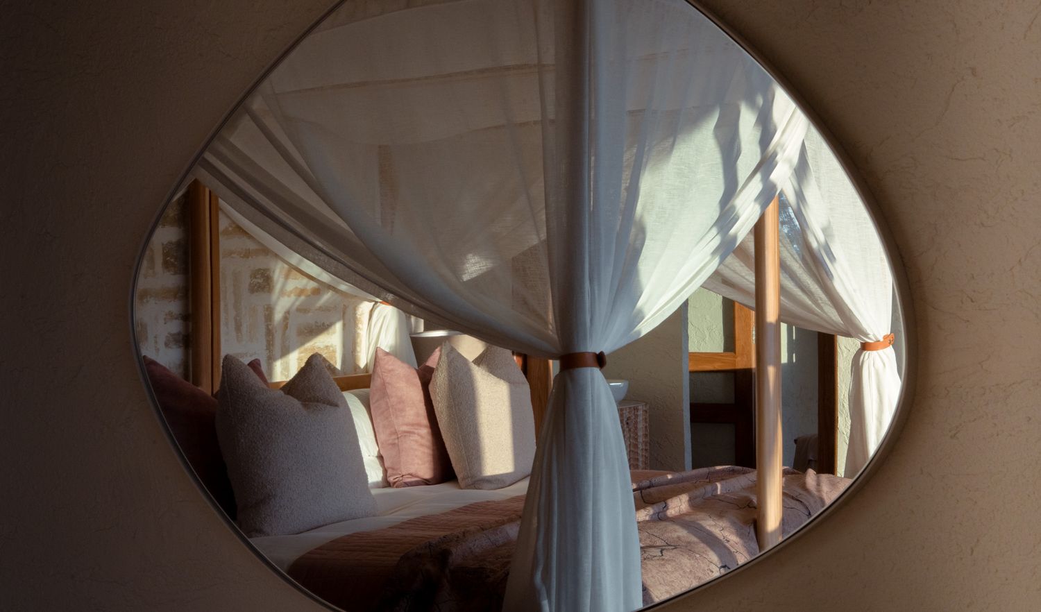 A cozy bedroom with white draped curtains and soft pillows viewed through an oval mirror.
