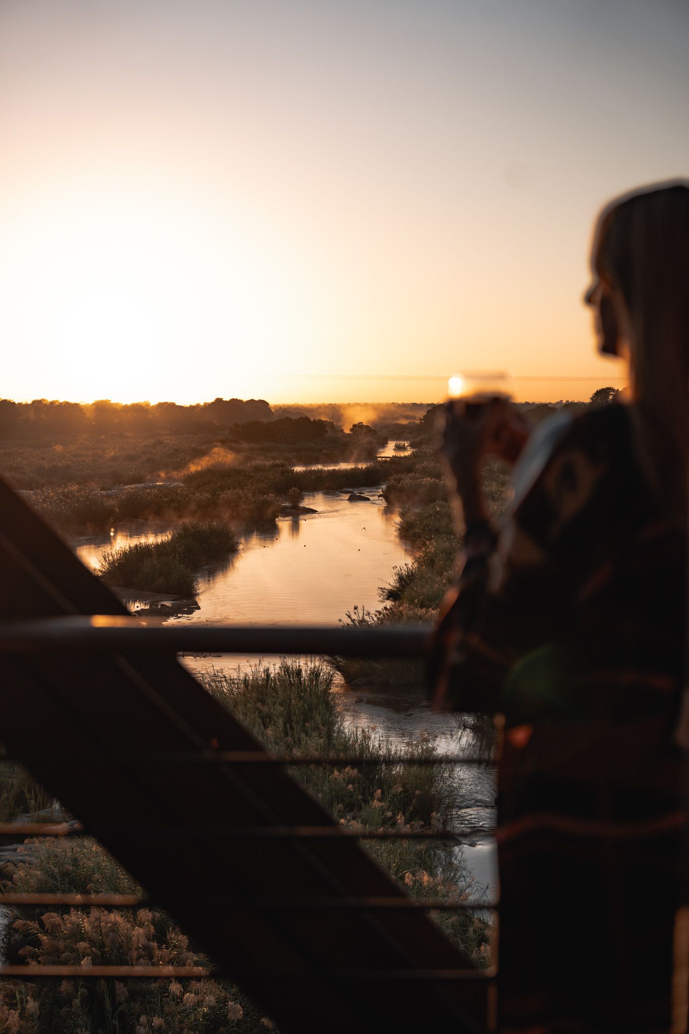Silhouette watching sunset over calm river with golden light reflecting off water and trees in distance.