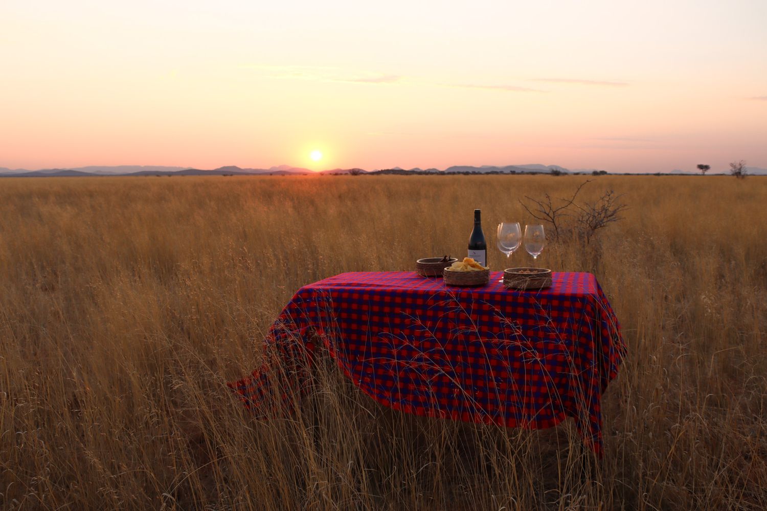 A romantic picnic setup with red tablecloth in an African savanna during sunset.