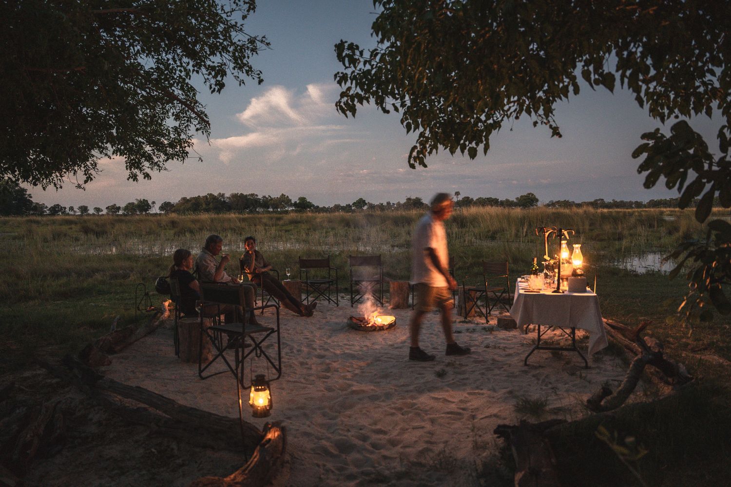 Evening campsite scene with campfire and camping chairs under trees.