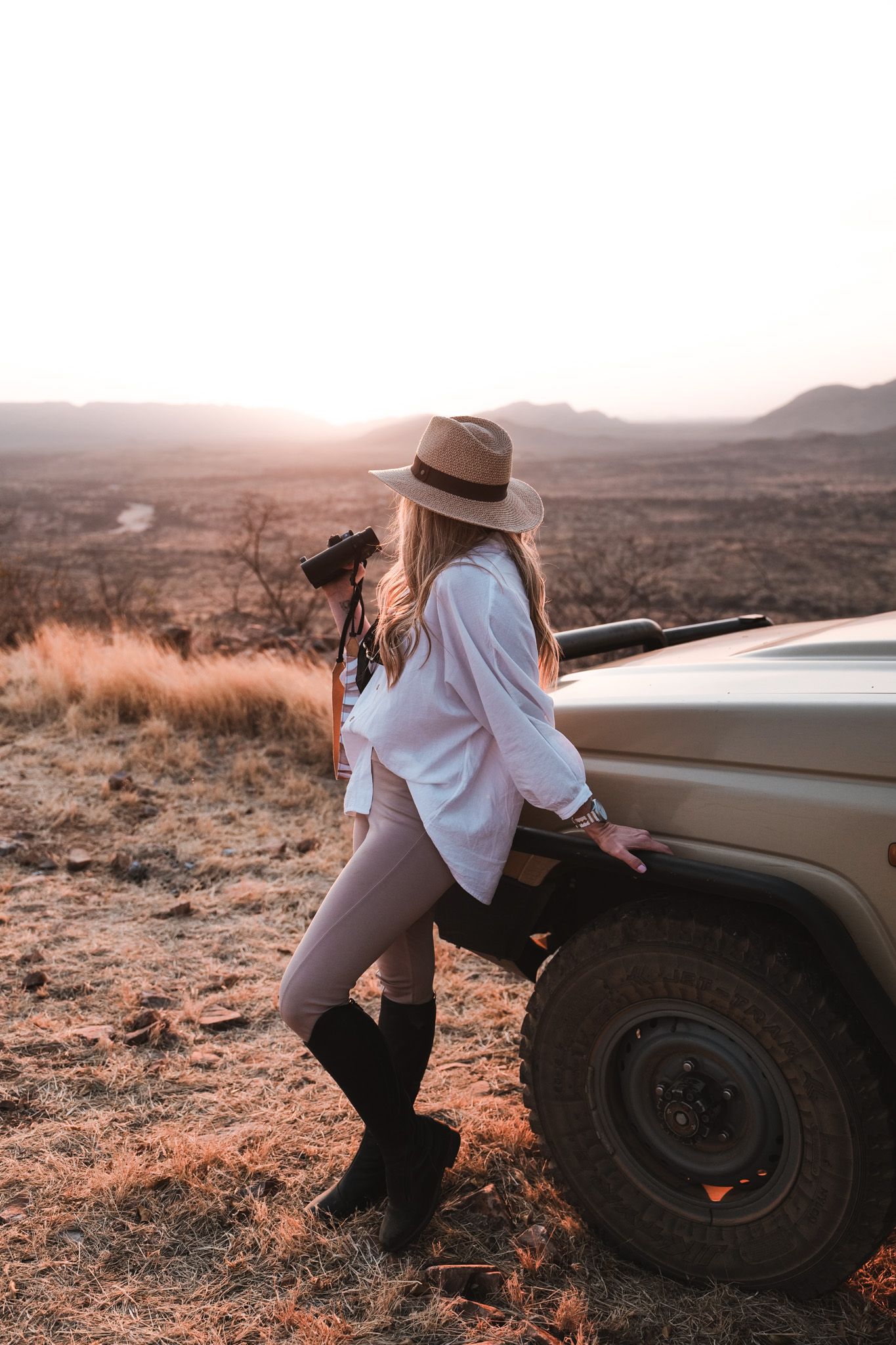 Adventure traveler in hat leans against safari vehicle in desert landscape at sunset.